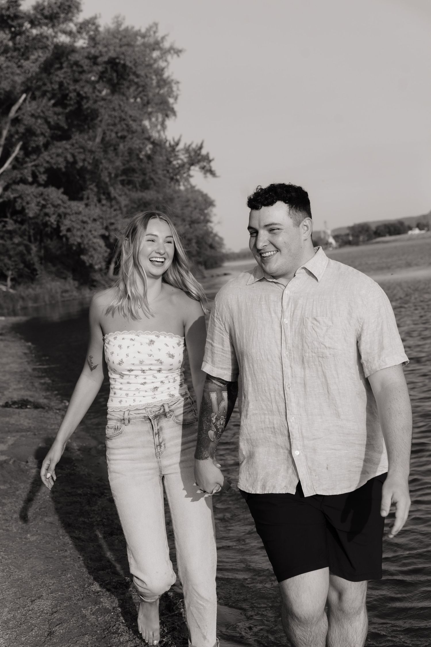 Black and white photo of a smiling couple holding hands and walking along a shallow shoreline with trees in the background.