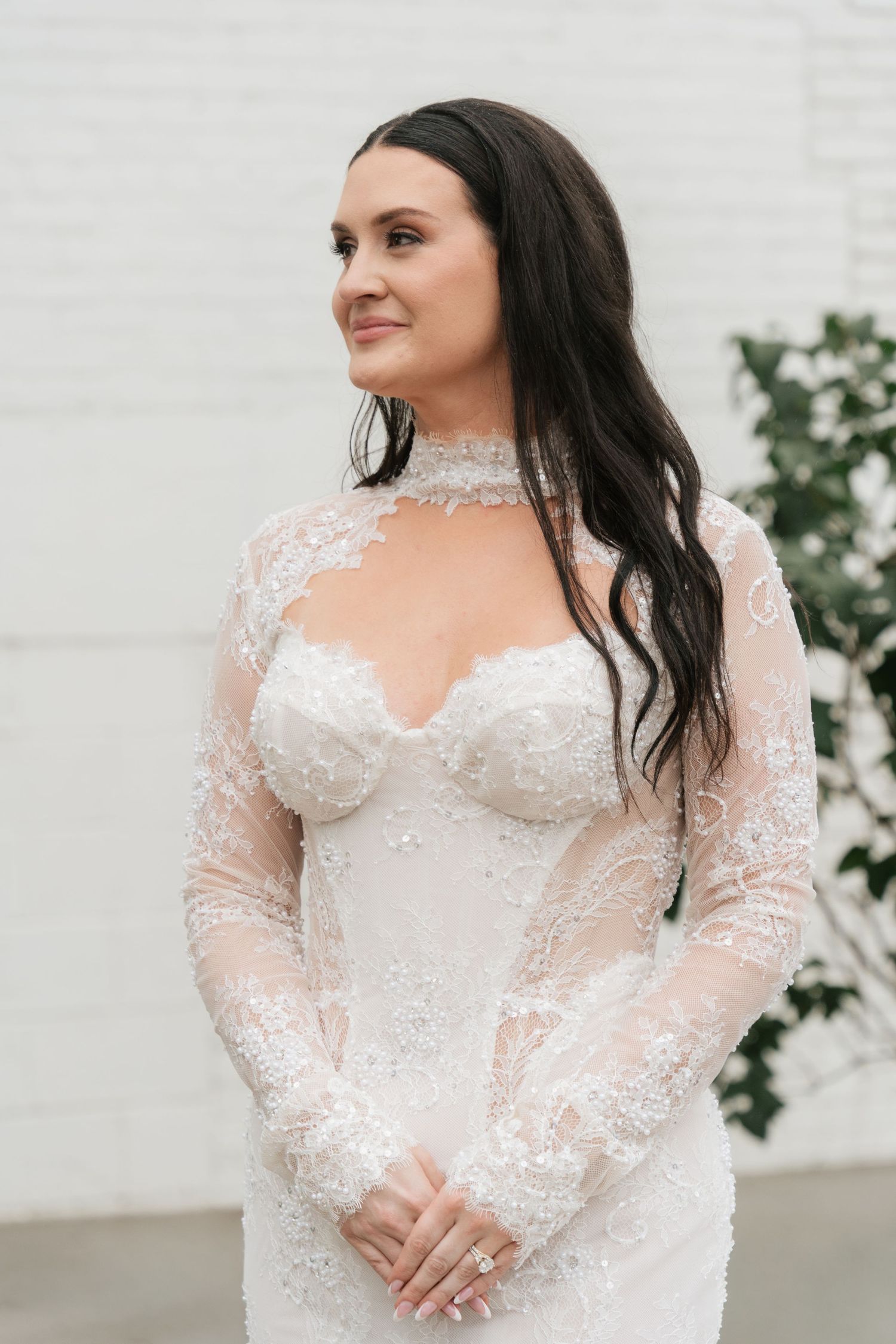 Bride in beaded lace long-sleeve wedding gown with sweetheart neckline and choker detail, posing against white brick wall.