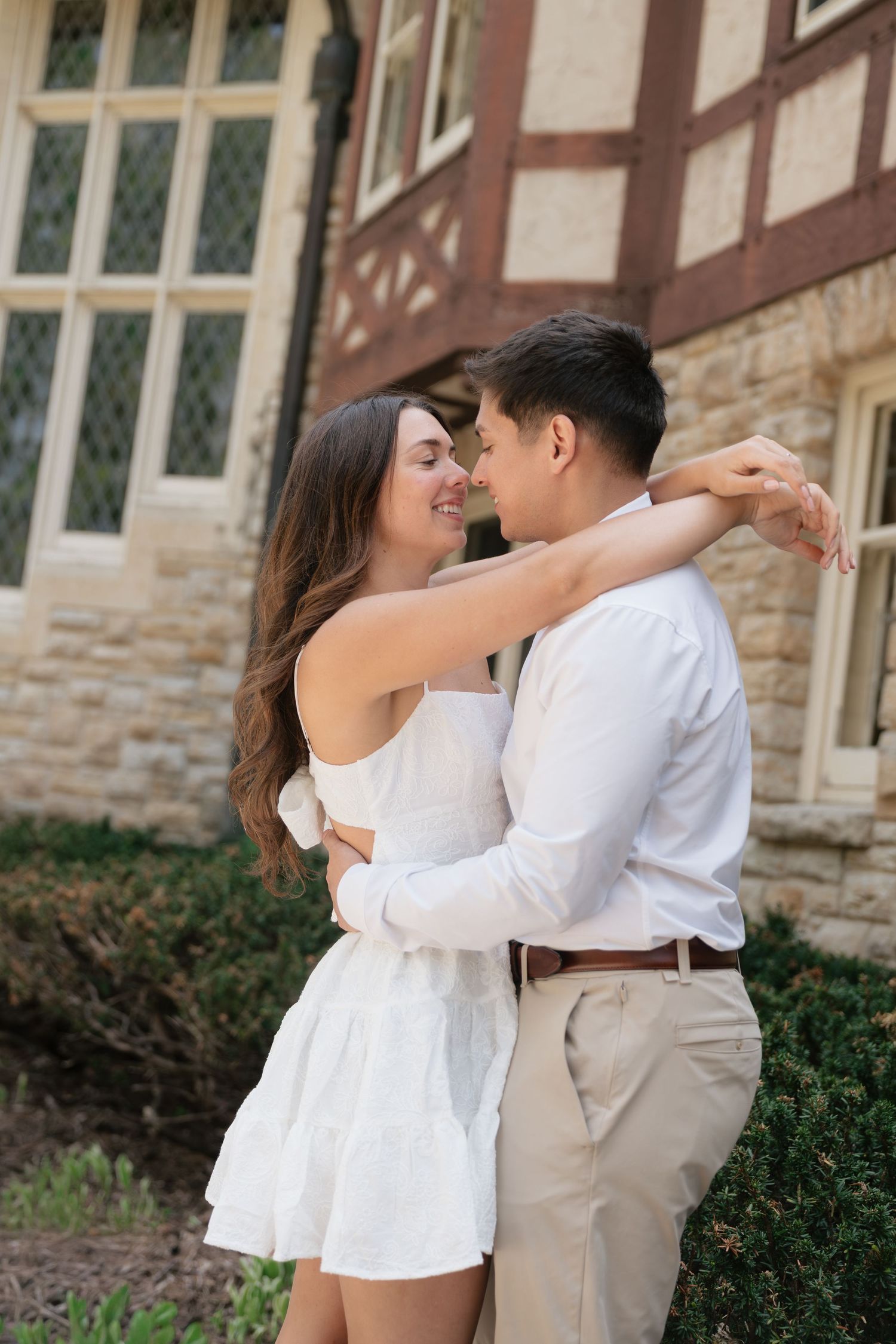 A couple in white outfits embrace and smile at each other outside a stone building surrounded by greenery.