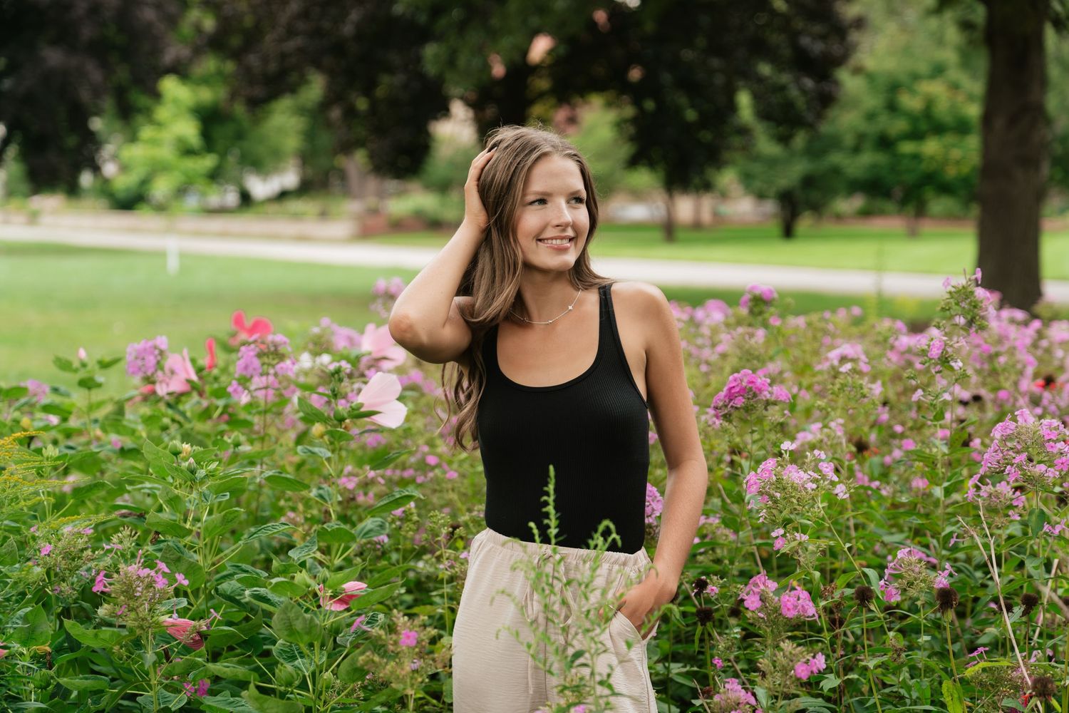 Smiling woman in black tank top standing among pink flowers in a lush green park on a sunny day.