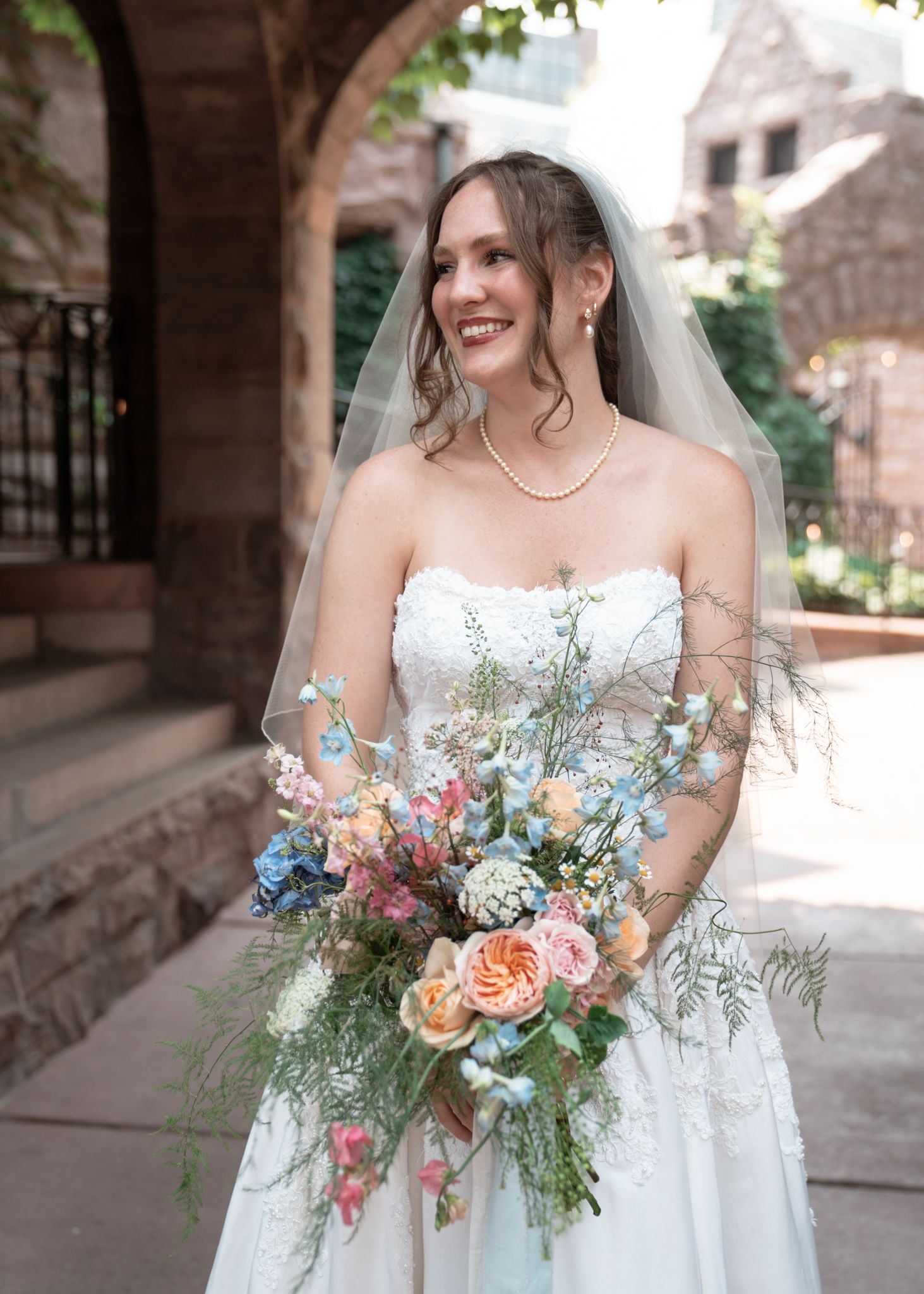 A bride in a strapless white gown holds a vibrant bouquet with blue and peach flowers in a stone archway setting. Van Dusen Mansion wedding.