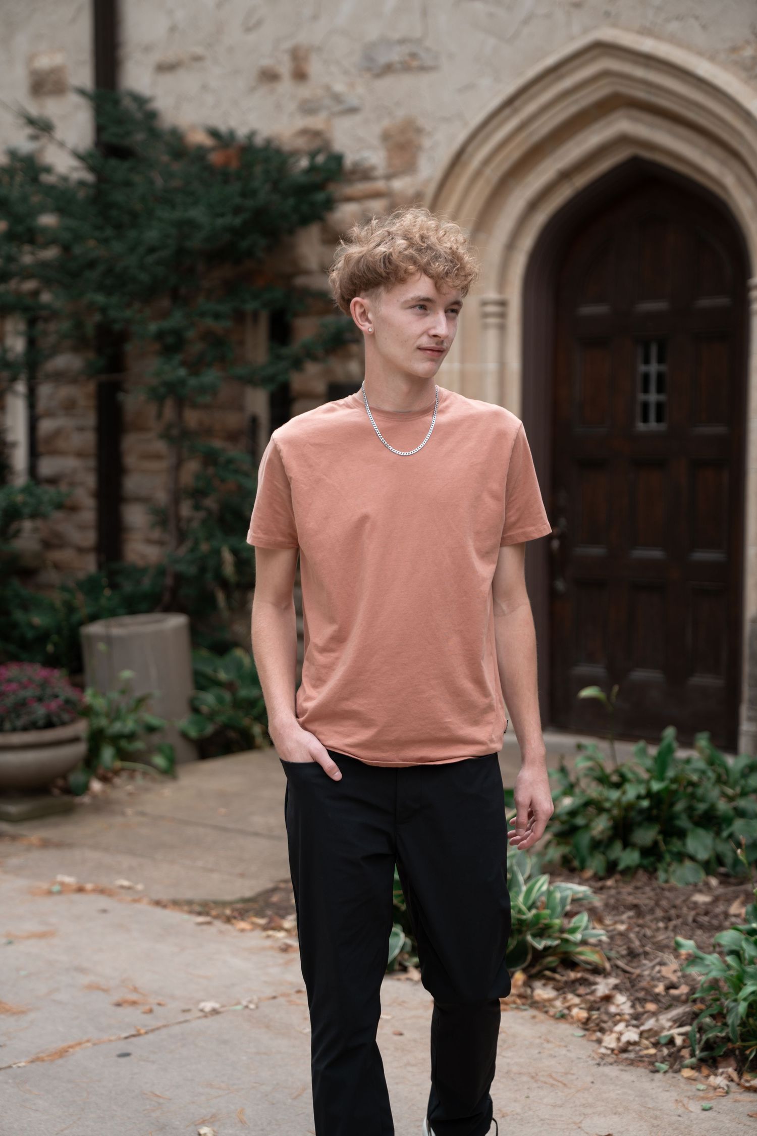 Person in salmon-colored shirt poses near gothic architecture and garden setting.