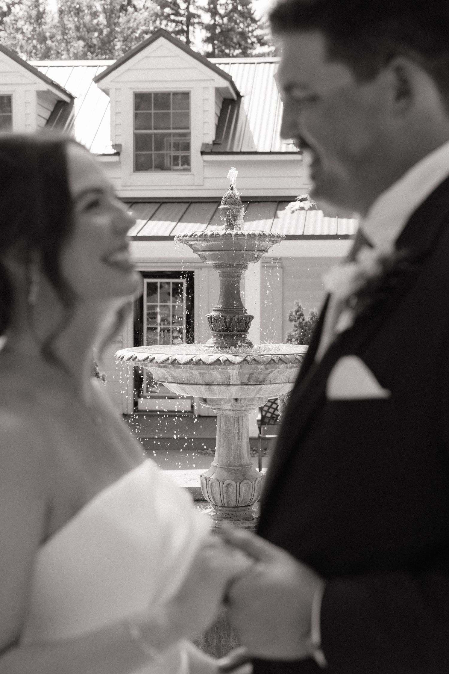 Black and white wedding photo of smiling bride and groom holding hands near elegant stone fountain outside historic venue.