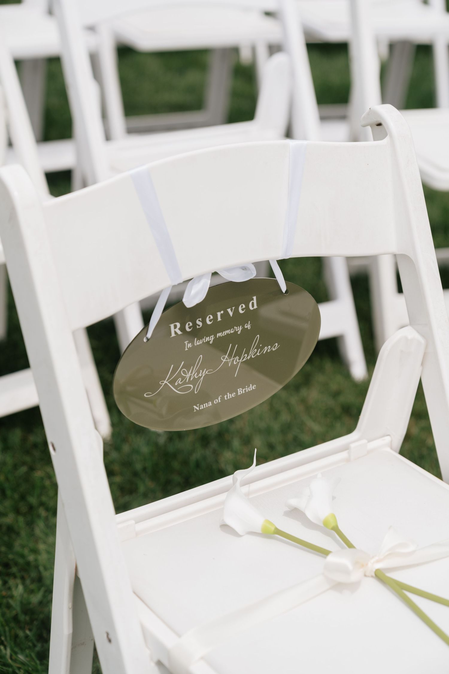 Reserved memorial chair sign for Kathy Hopkins, Nana of the Bride, with white calla lilies at an outdoor wedding.
