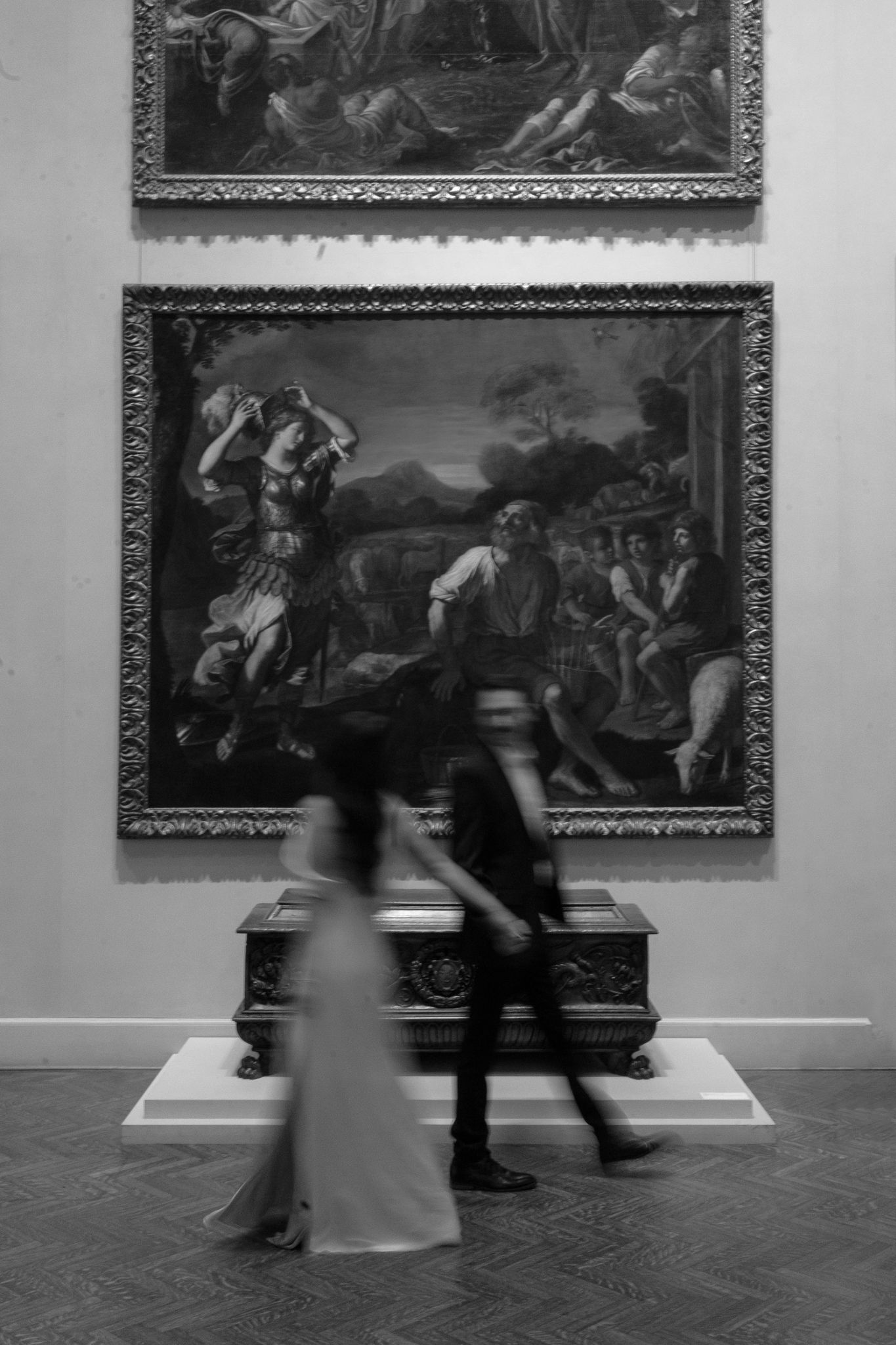 Art gallery interior with classical paintings displayed on white walls as visitors pass through.