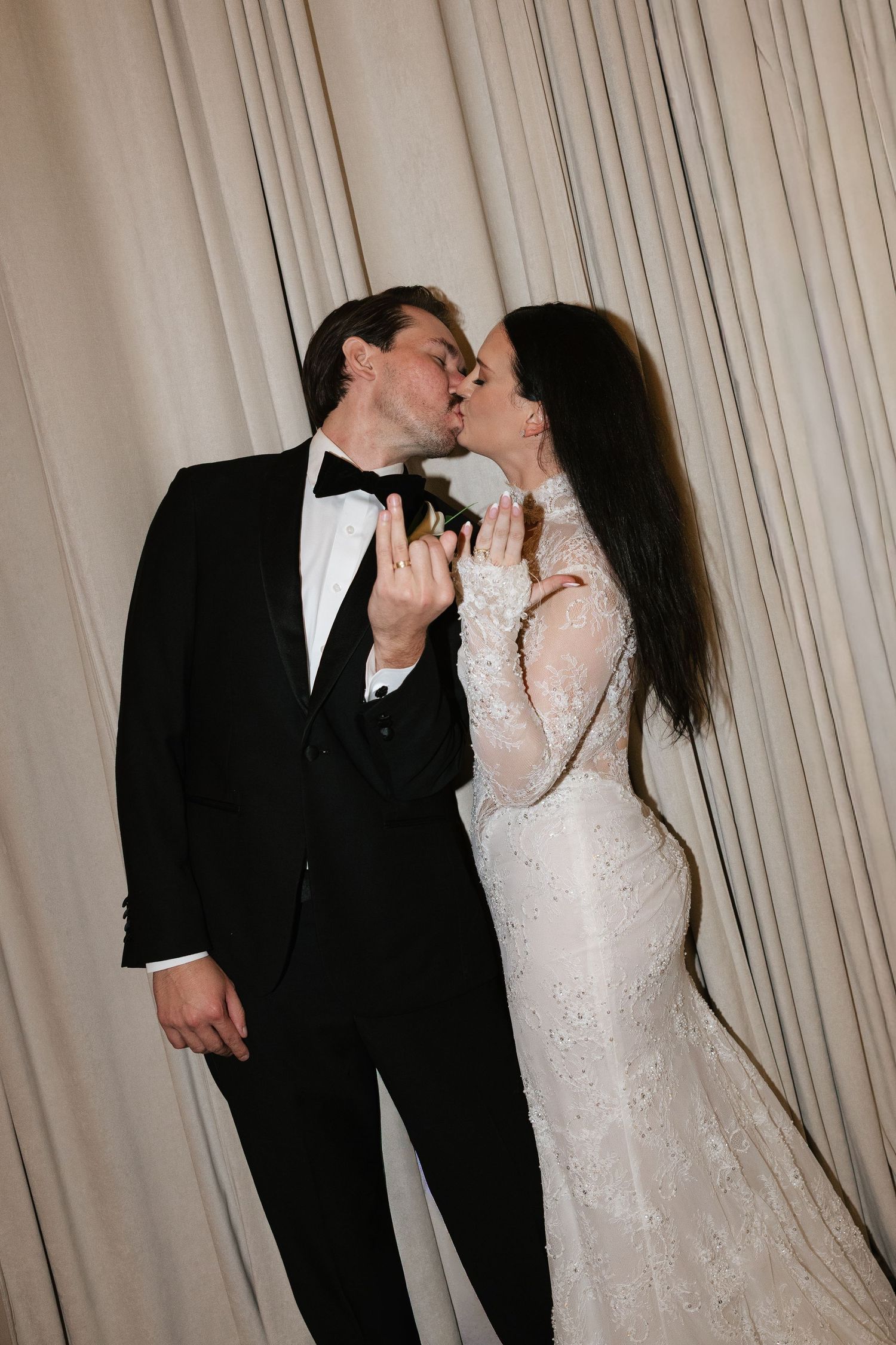 Bride and groom sharing a kiss in wedding attire, she in lace gown, he in black tuxedo with bow tie.