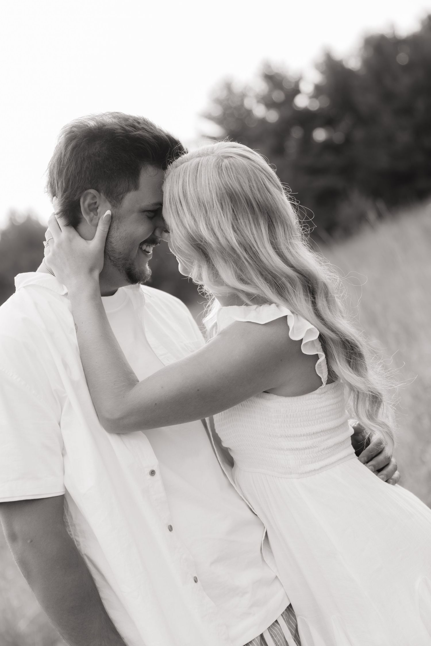 Black and white photo of a romantic couple in white outfits embracing and smiling at each other in a field.