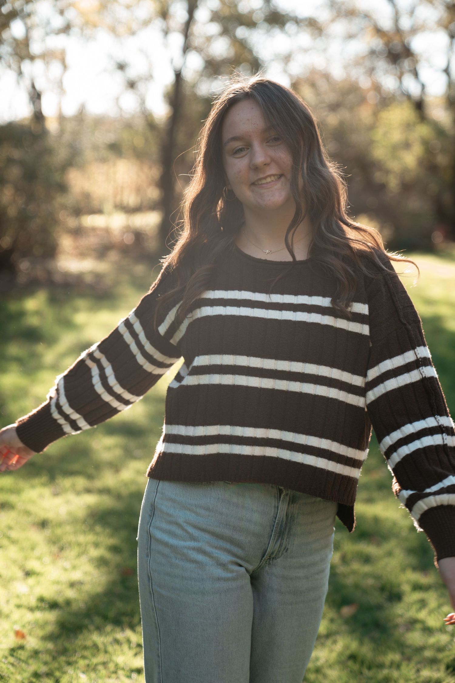 A casual outdoor photo showing someone in a brown and white striped sweater and light jeans against a sunny natural backdrop.