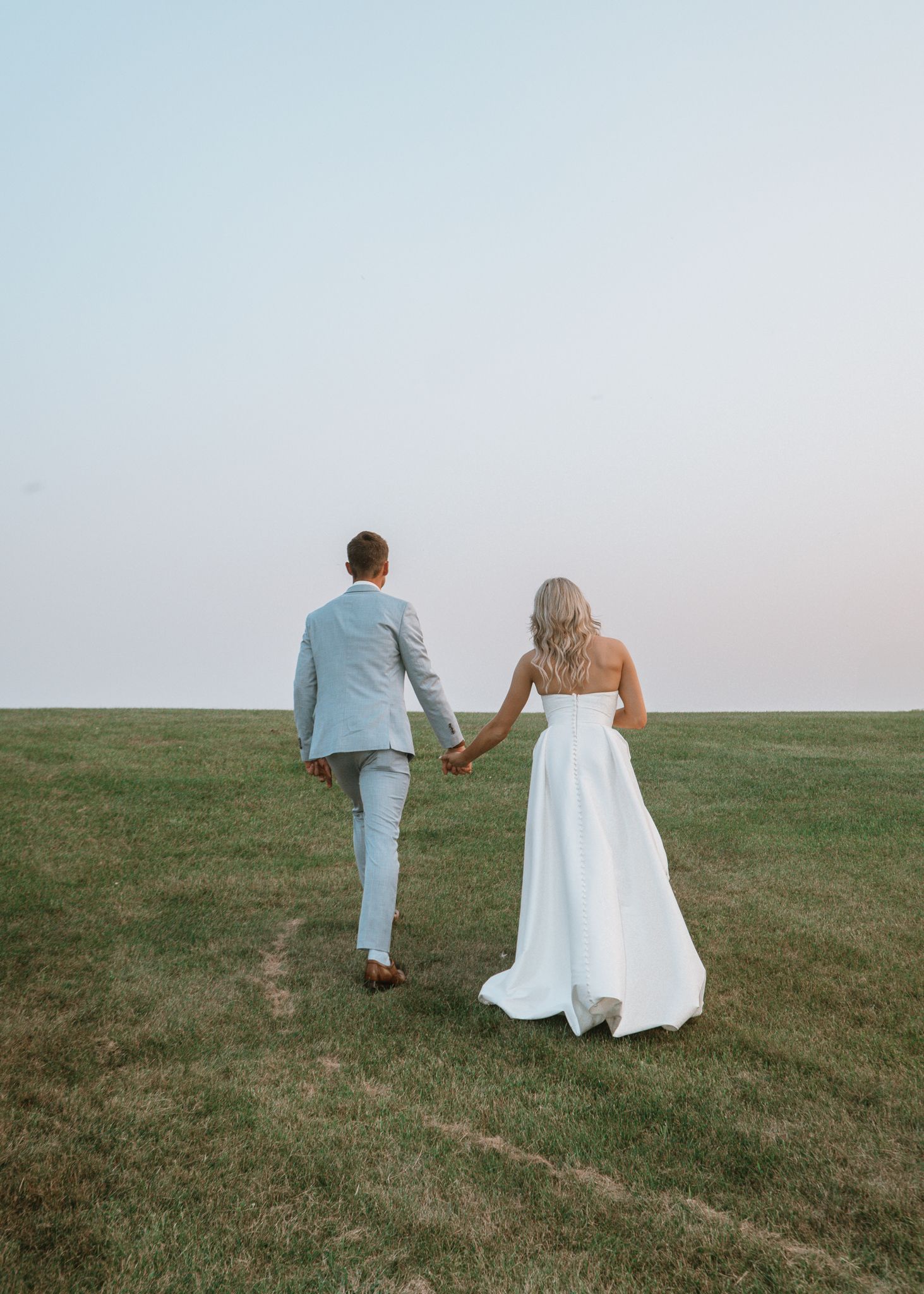 A couple in wedding attire walks up a grassy hill at sunset holding hands.