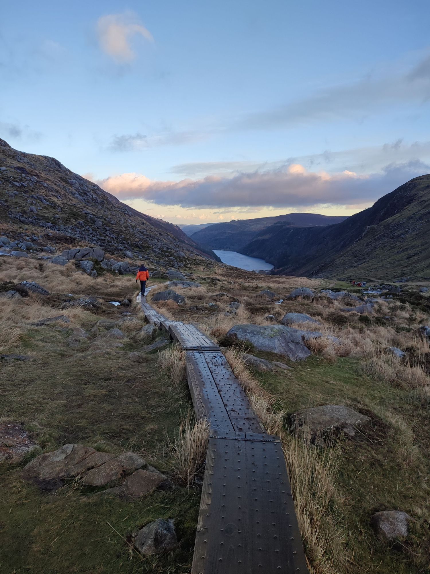 Wooden boardwalk path leading the scenic mountain views and Glendalough at sunset with a lake in the distance.
