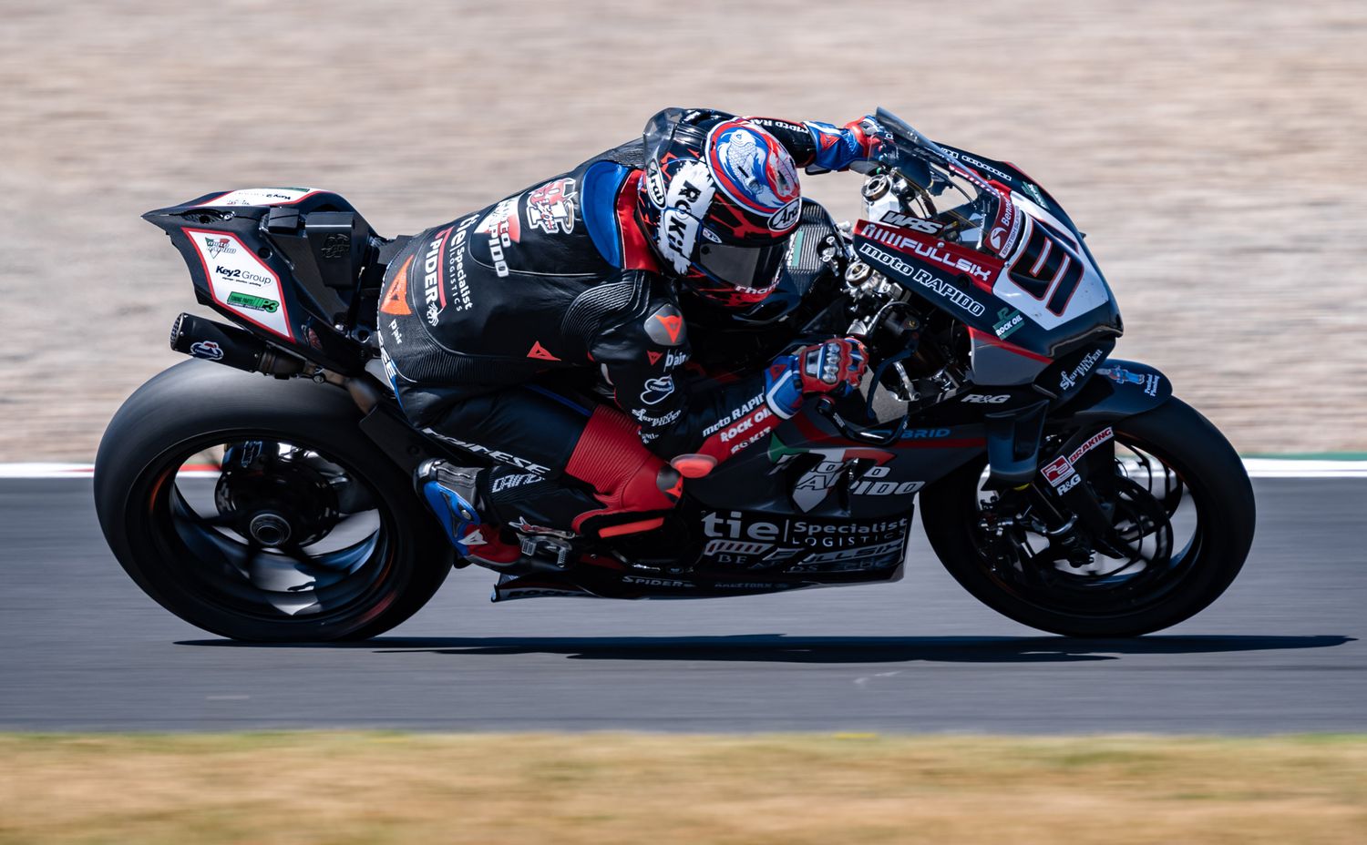A motorcycle racer leans into a turn on a racing circuit, riding a black and blue sportbike at high speed.