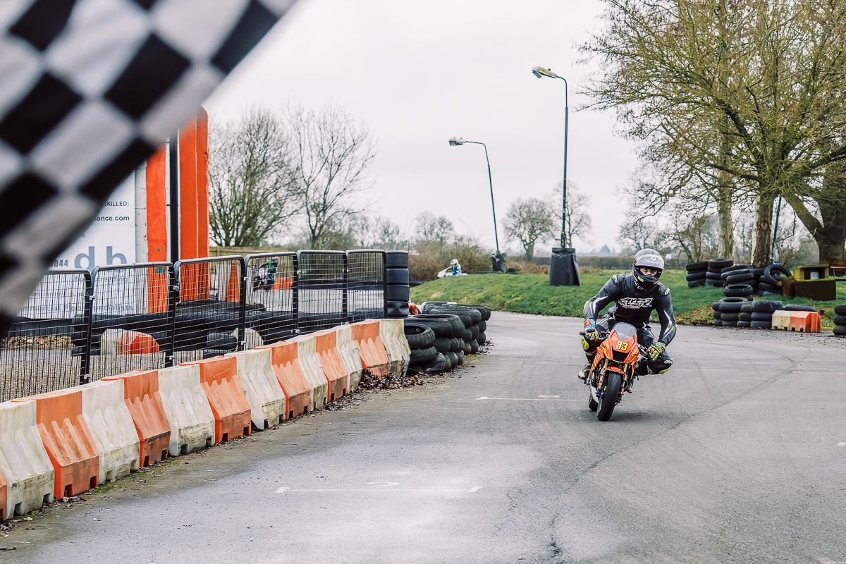 Racing motorcycles navigate track with orange barriers and checkered flags visible.