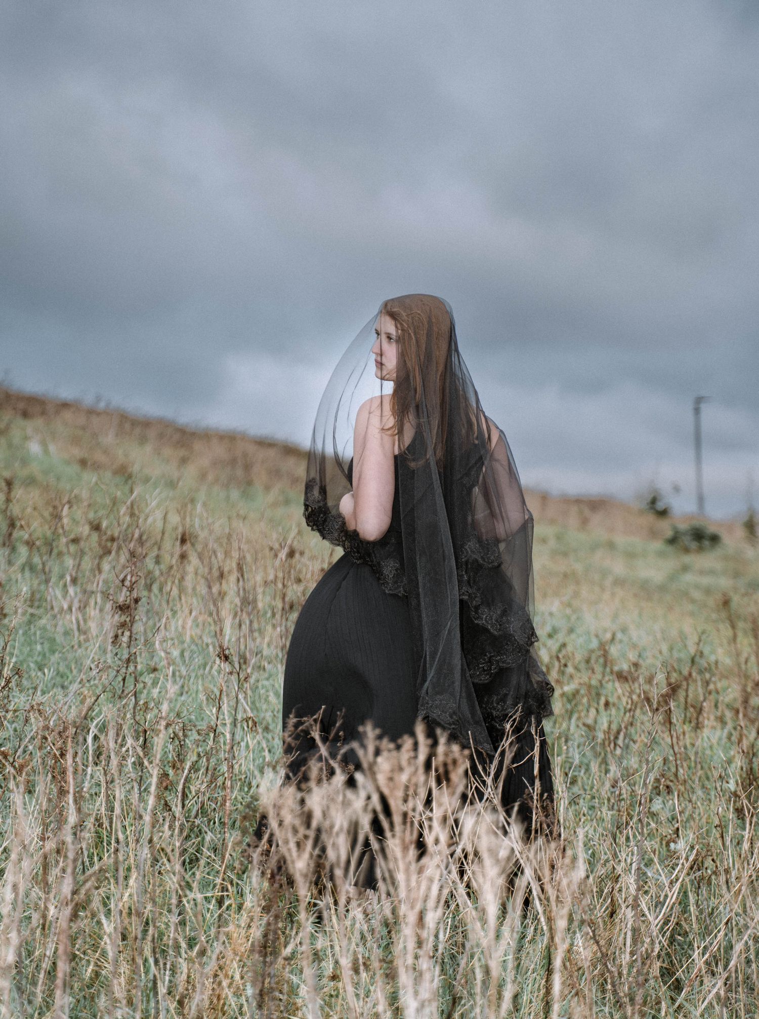 Person in dark dress standing in tall grass on St Ives cliffs during a Cornwall elopement under moody storm clouds