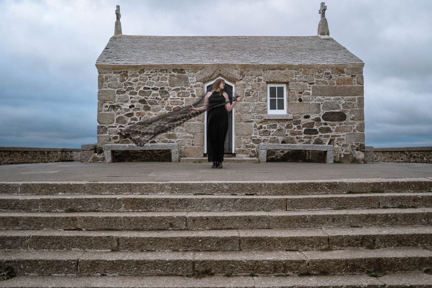 Bride standing on steps outside stone chapel in Cornwall gothic wedding setting