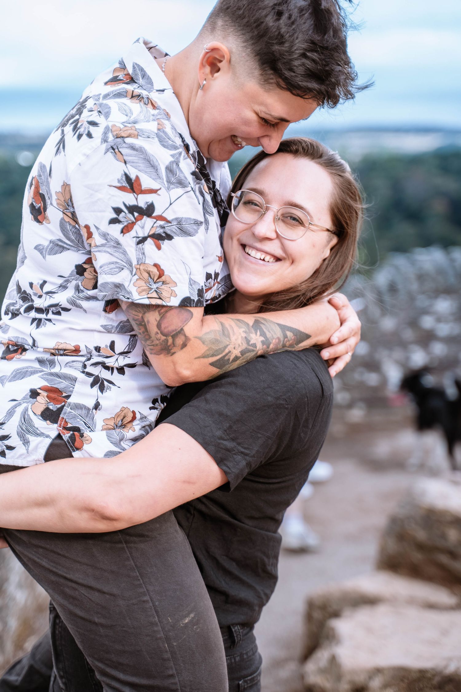 A same-sex couple share a playful lifted hug at Symonds Yat Rock.