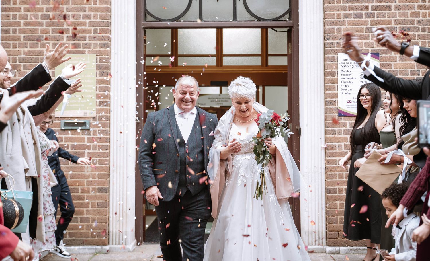 Joyful wedding party celebrates outside brick building with confetti flying through the air.