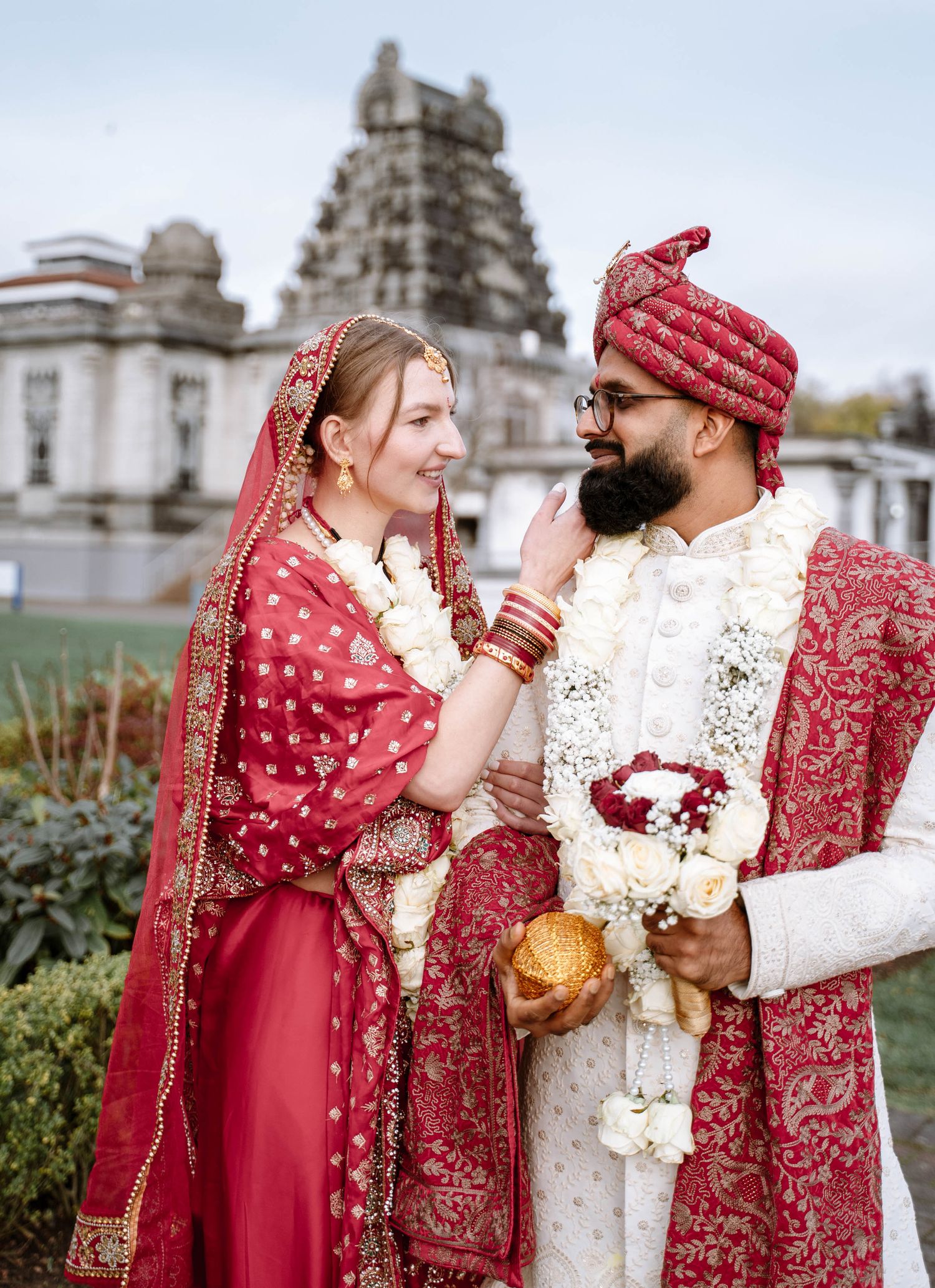 A couple in traditional wedding attire share joyful moments in front of ornate architecture, wearing red and cream garments.