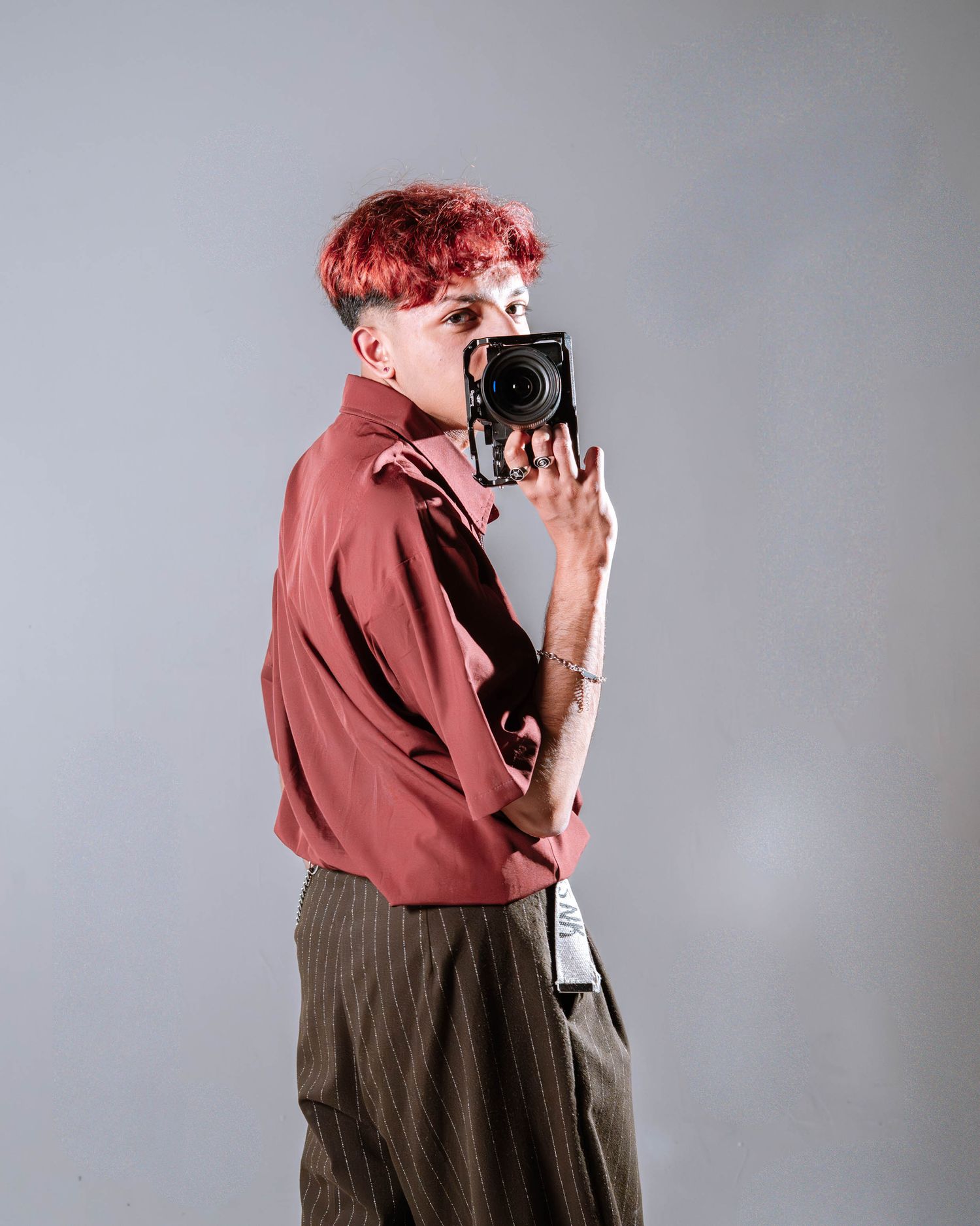 Person with pink hair in a red shirt holds a camera up to their face against a gray background.