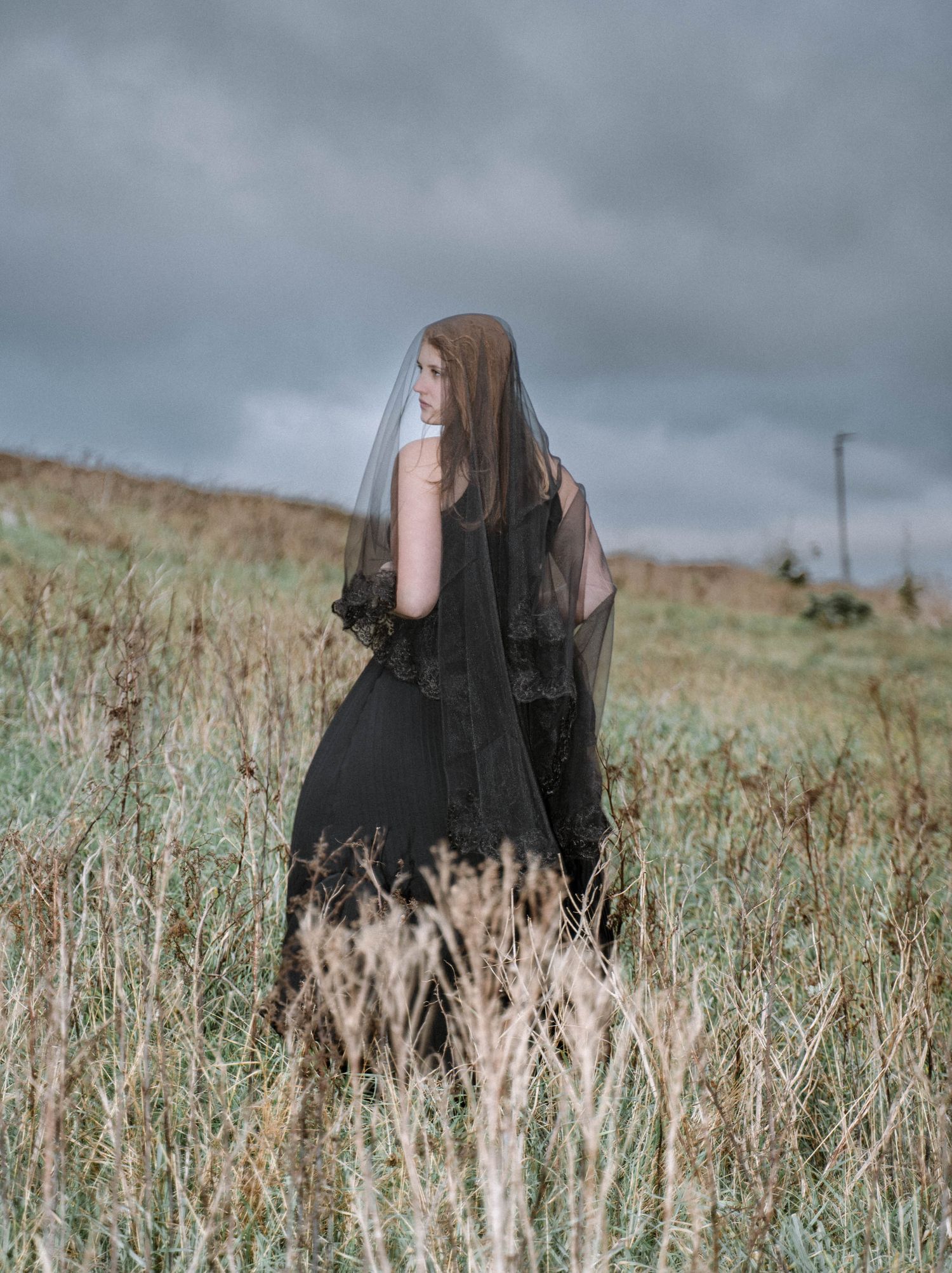 Cornwall gothic bridal portrait woman in black dress walking through field moody editorial elopement photography Cornwall UK