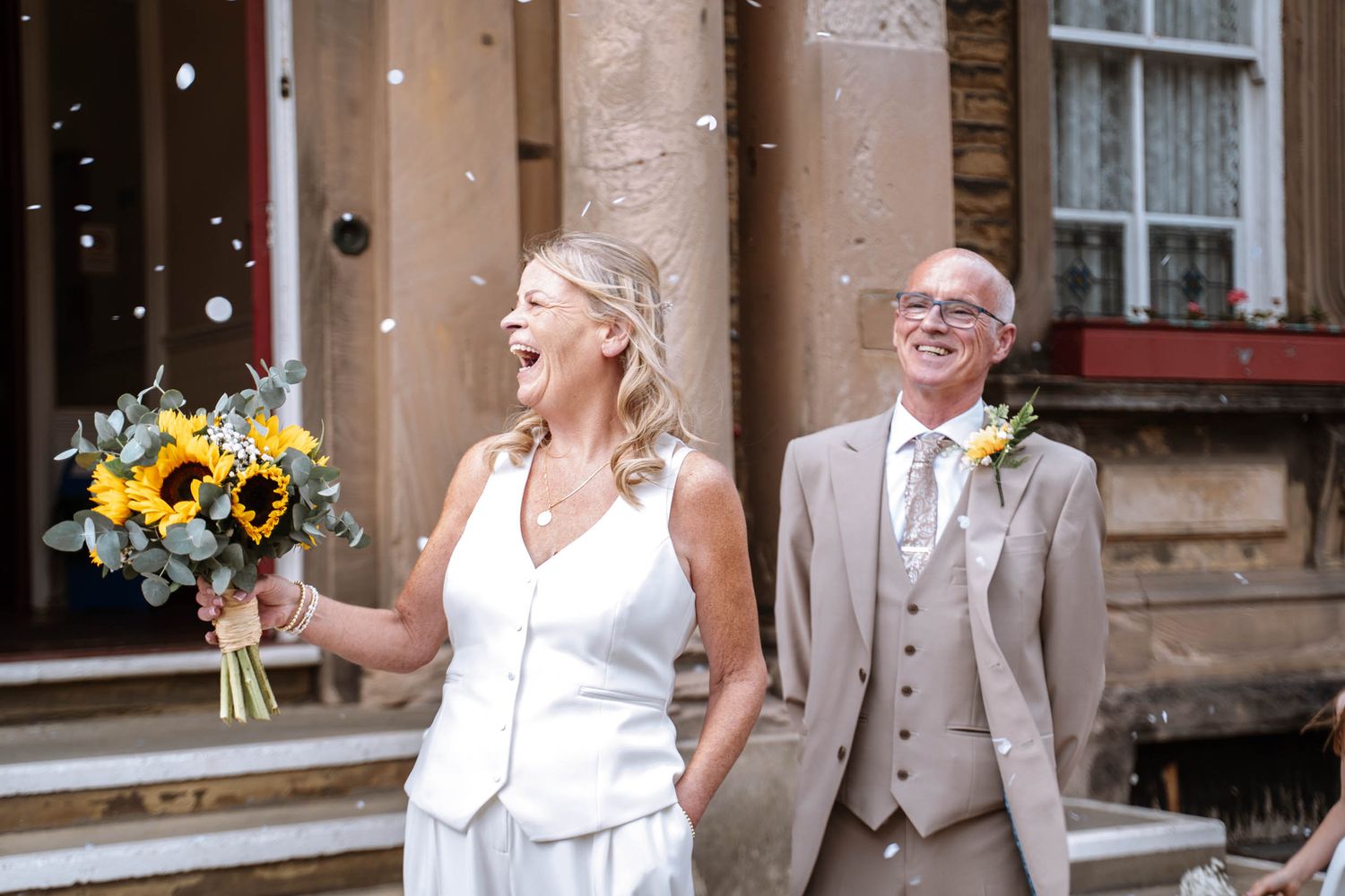 Bride laughing with a sunflower bouquet as confetti falls outside Waterloo Town Hall, groom beside her in a beige suit