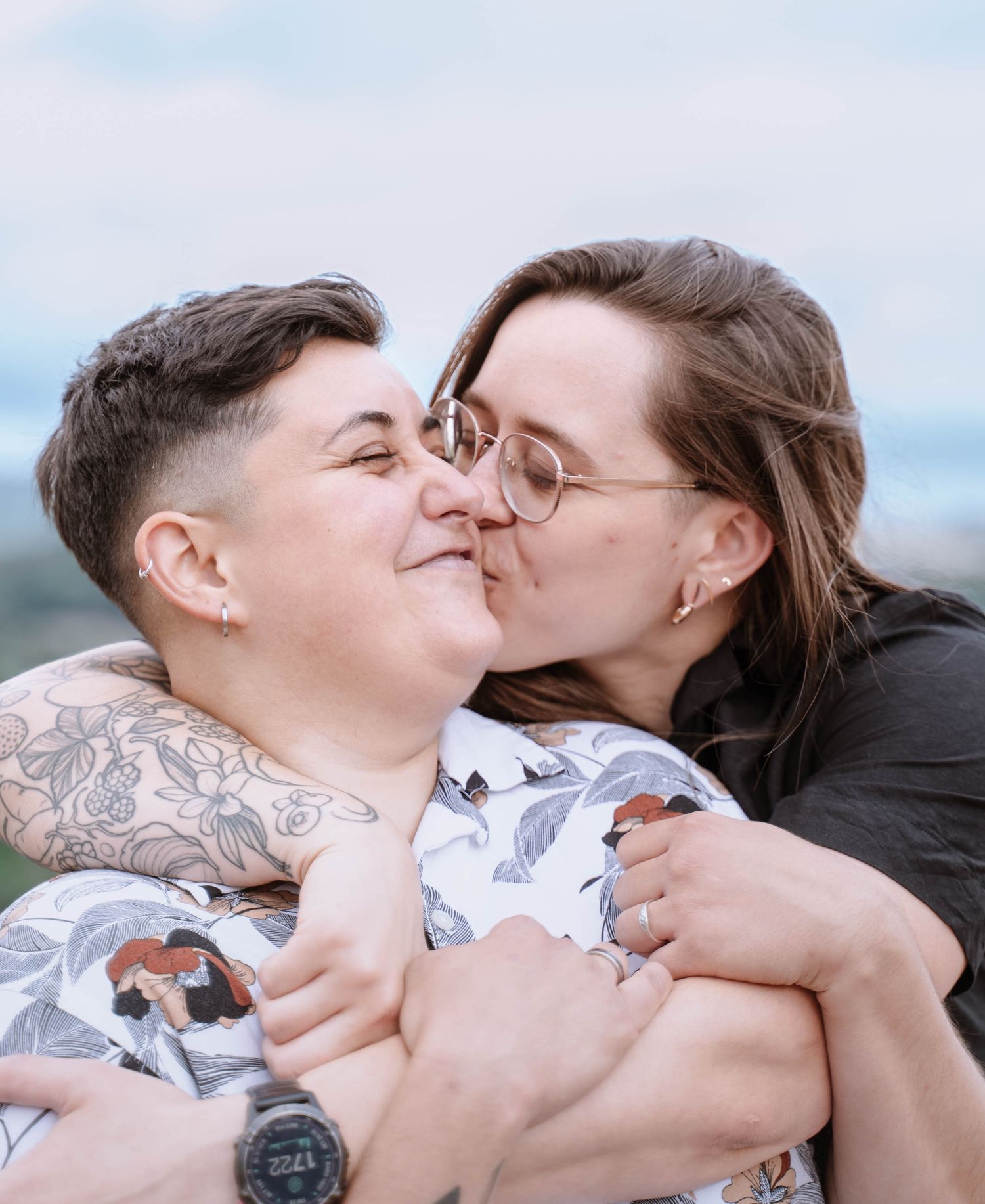 A queer couple smile as one kisses the other on the cheek after their proposal.
