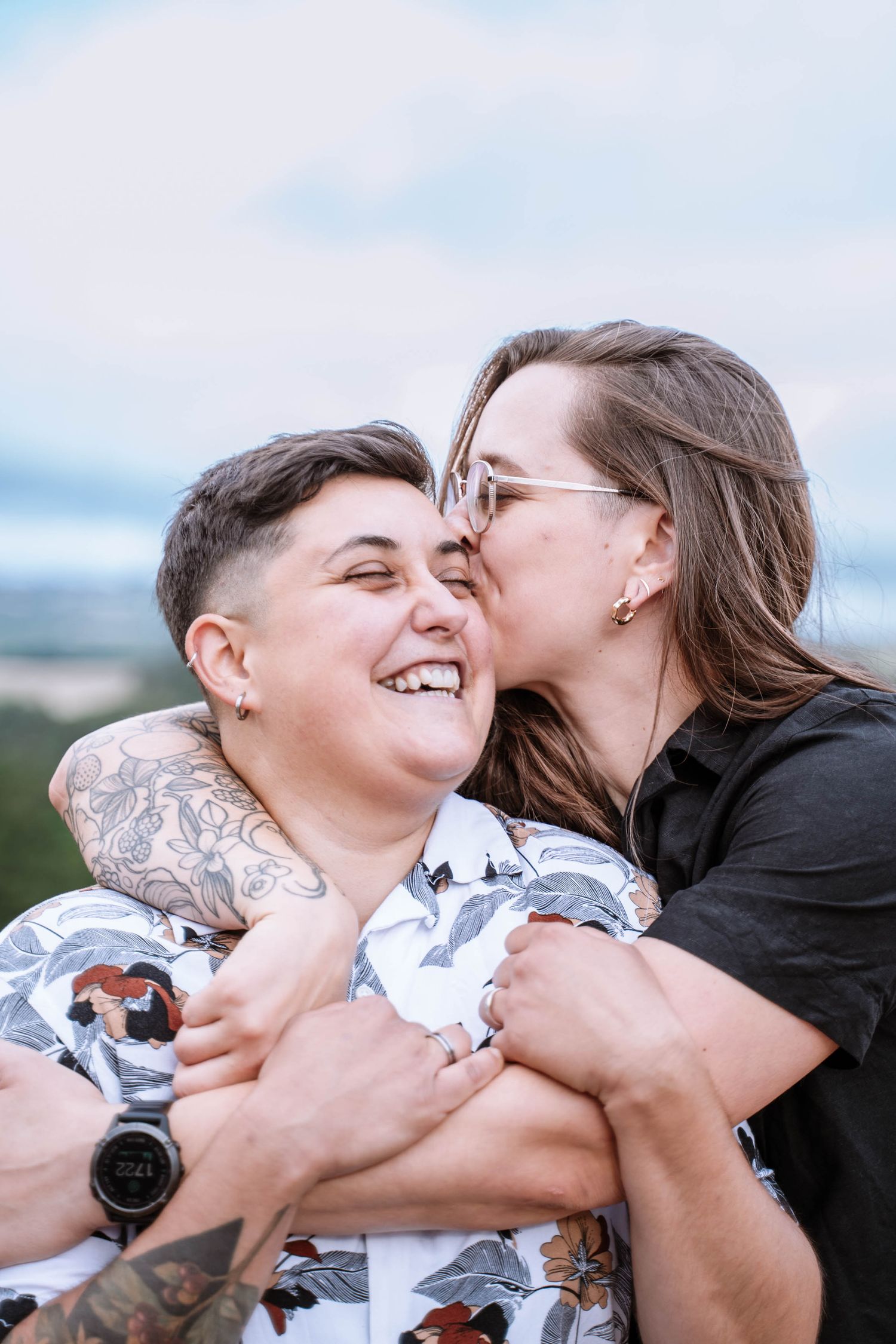 A queer couple laugh as one kisses the other’s temple during a close portrait.