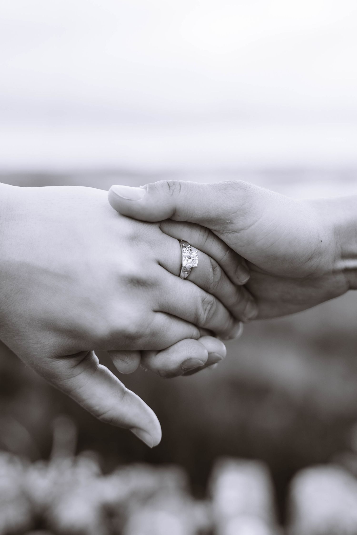 Black-and-white close-up of a queer couple holding hands with an engagement ring visible.
