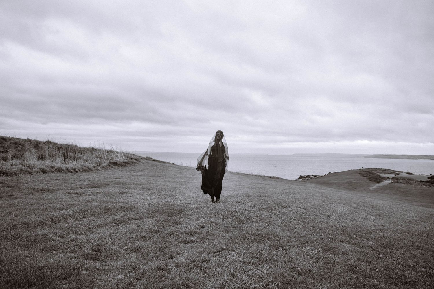 Lone figure in black veil walking across coastal field during a St Ives elopement with wide cinematic Cornwall landscape