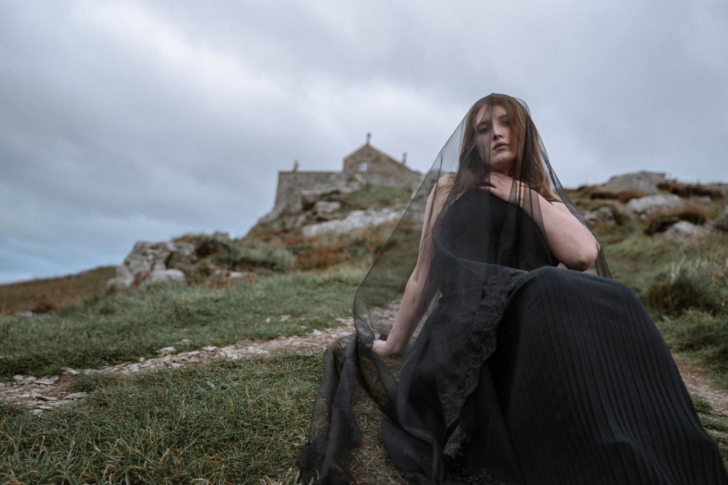 Bride posing in black dress near cliffside chapel in Cornwall