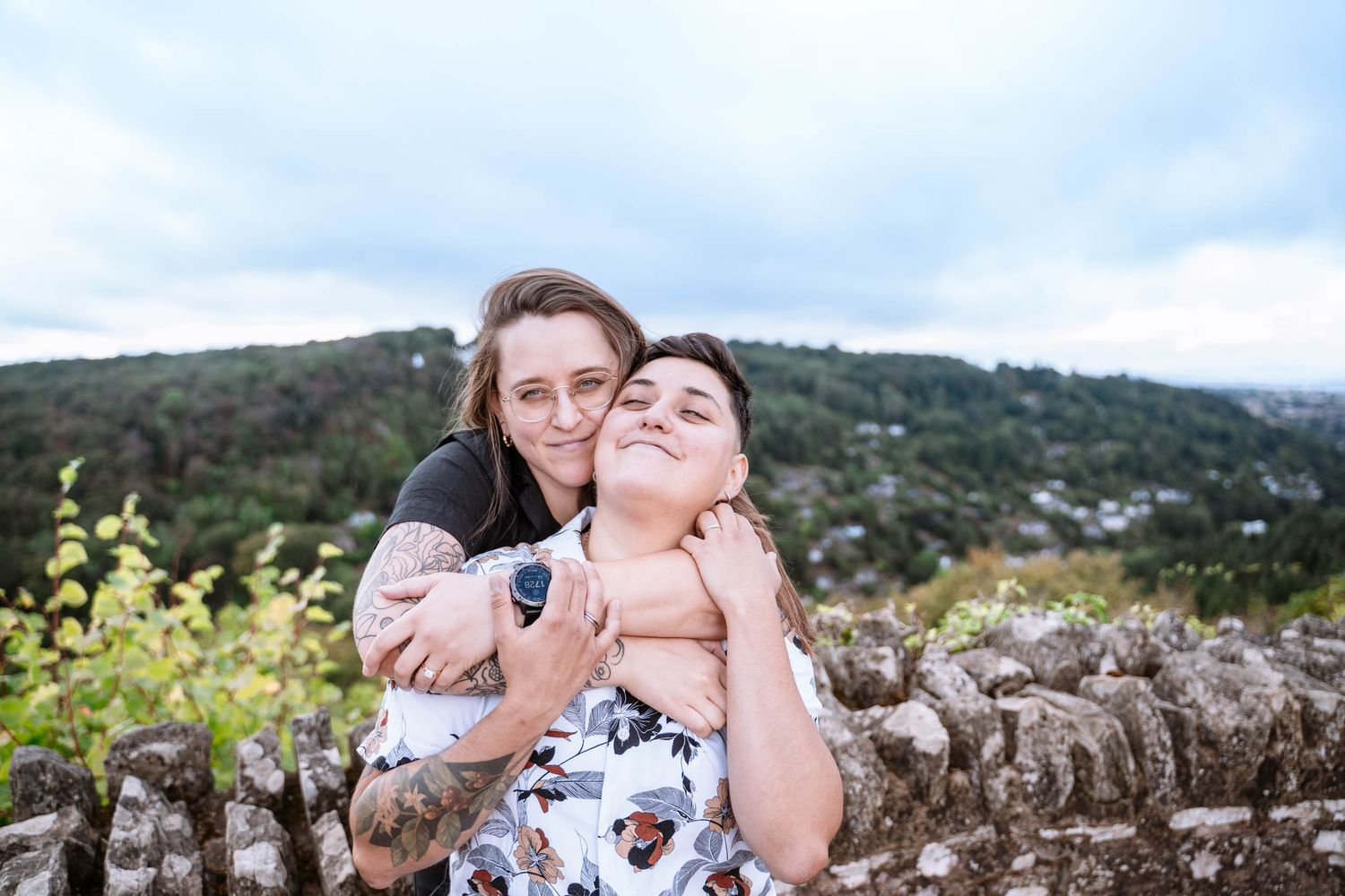 A newly engaged lesbian couple share a playful back hug beside the stone viewpoint wall.