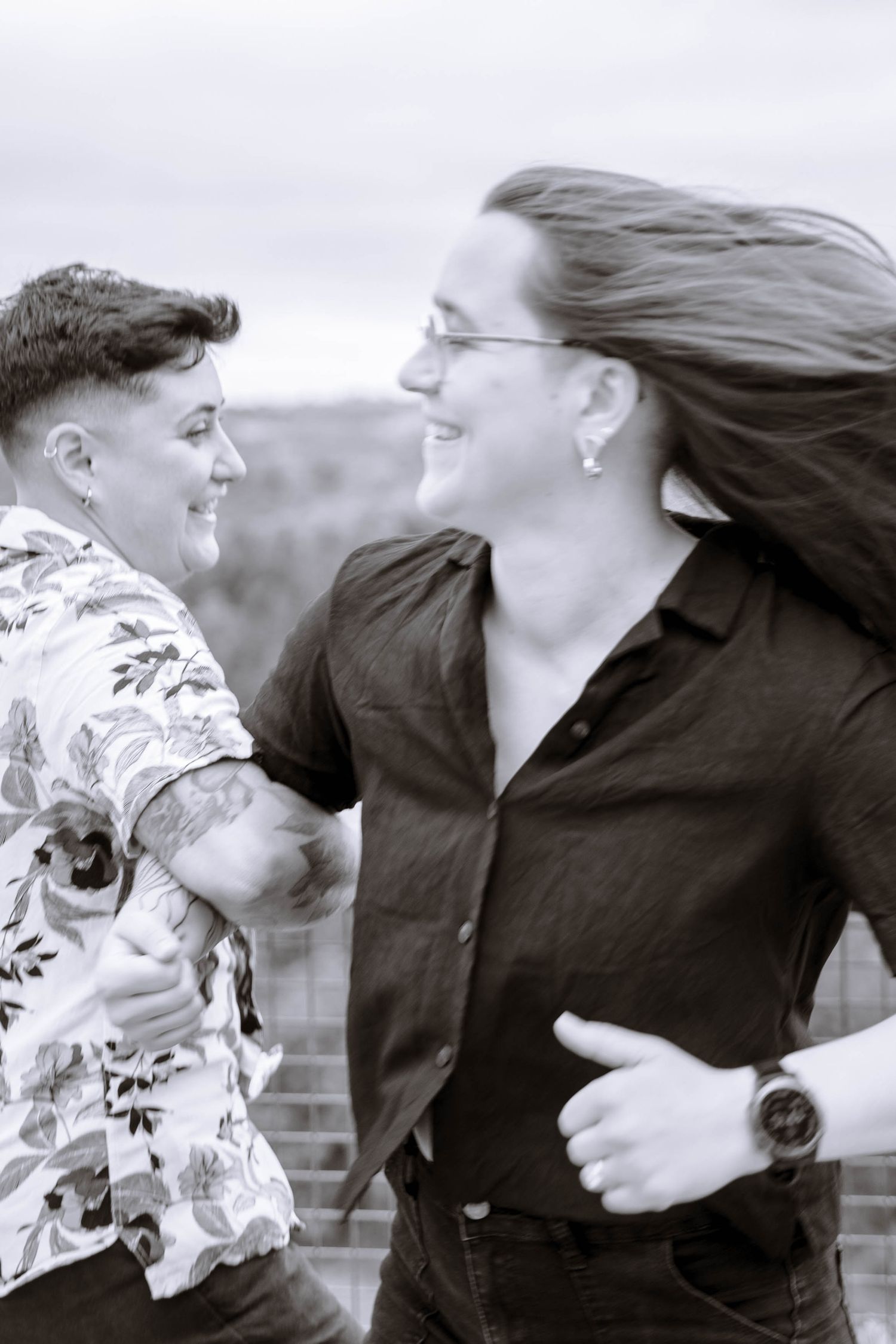 Black-and-white candid portrait of a lesbian couple laughing together in the wind.