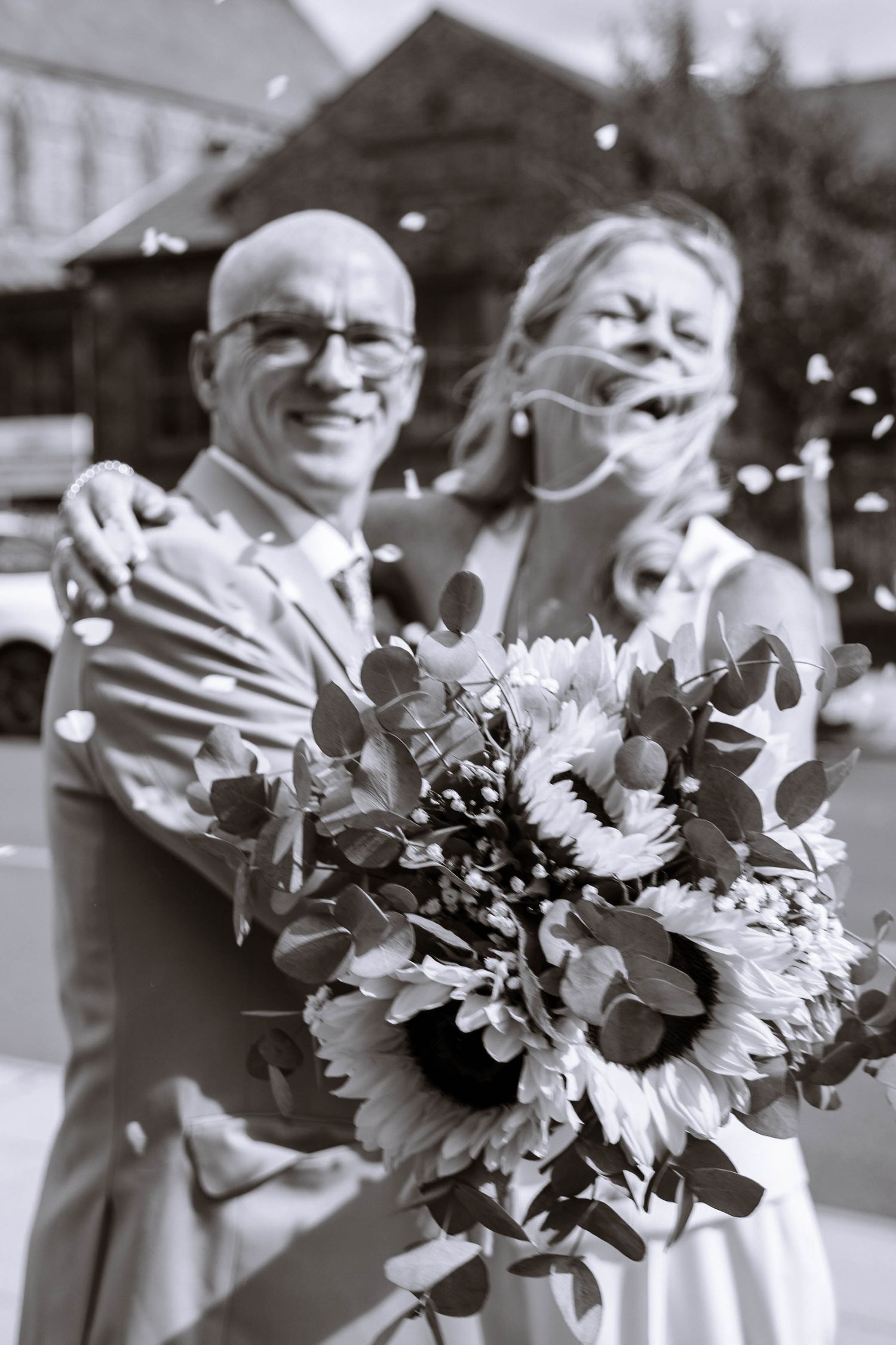 Black-and-white portrait of the bride and groom laughing together with a bouquet as confetti falls outside Waterloo Town Hall