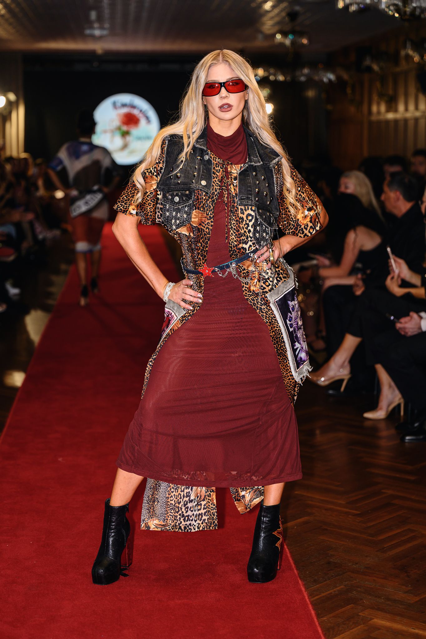 Model poses in eclectic mixed-pattern outfit with red sunglasses on runway.