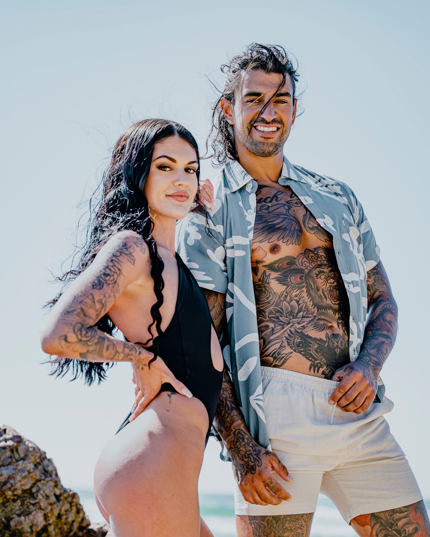 Couple in beachwear poses together against bright sky background.