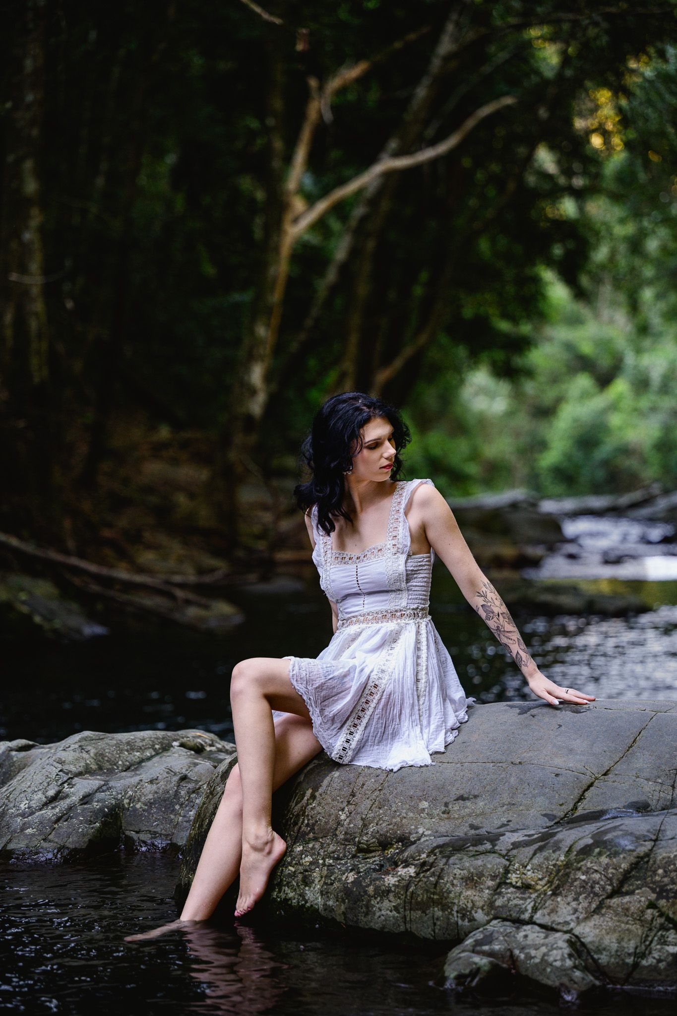 Woman in white lace dress sitting on rocks by a forest stream, with tattoos and bare feet touching the water.