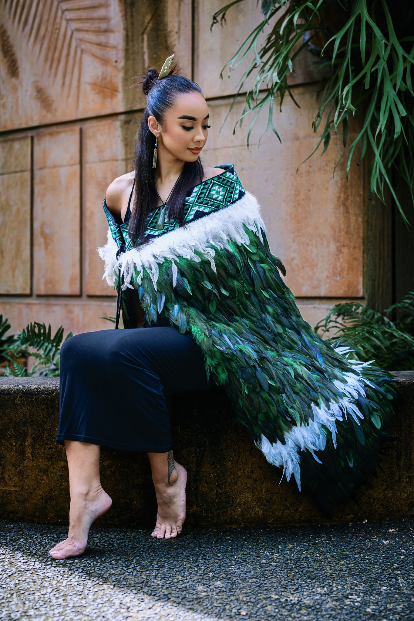 Woman wearing traditional Maori korowai feather cloak in green and white, seated barefoot outdoors.