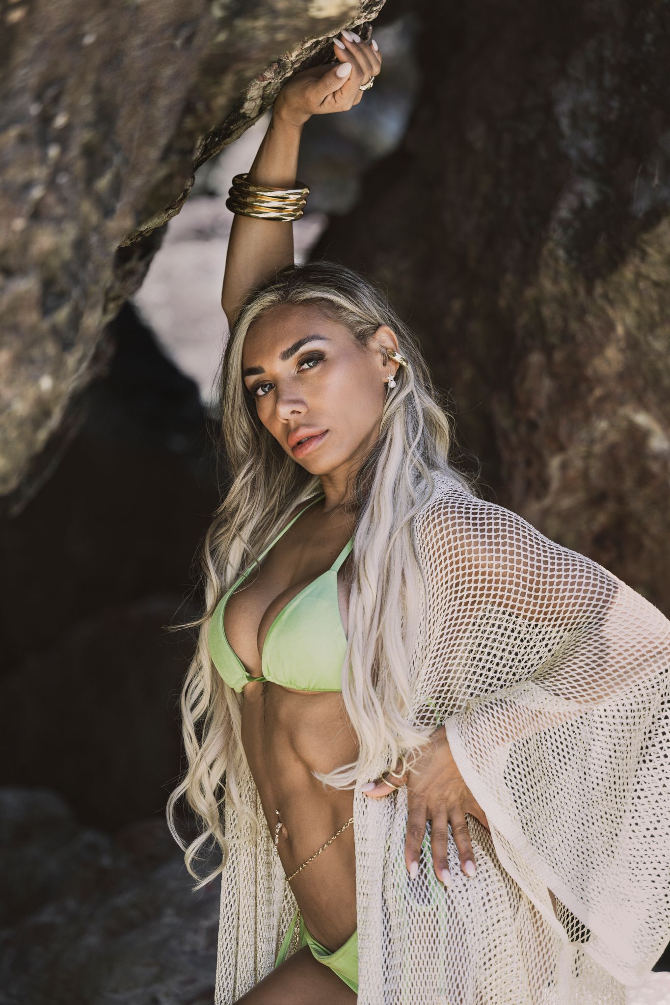 Model in green swimwear and mesh cover-up poses with raised arm against cave wall.
