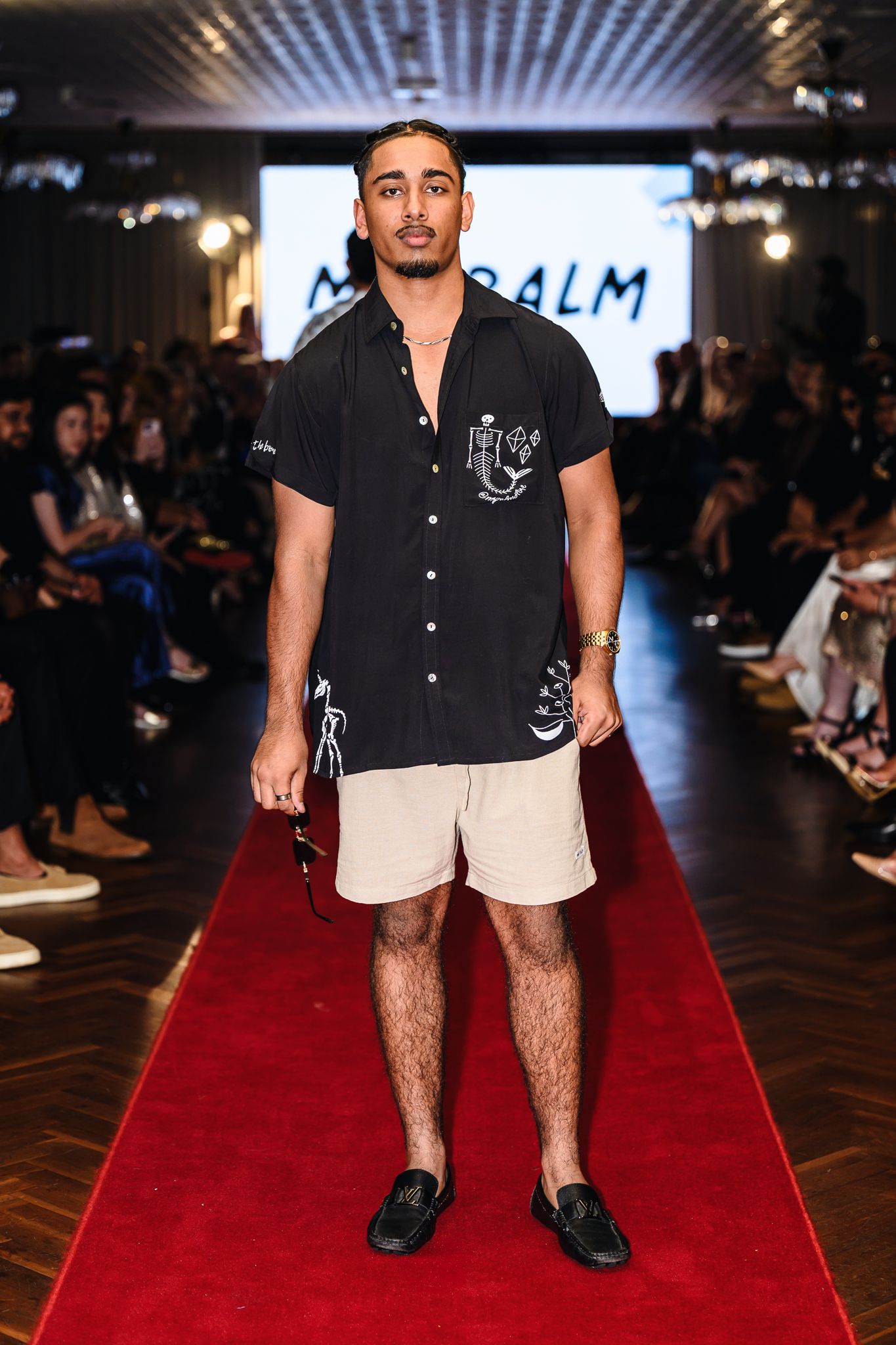Model wearing black shirt and khaki shorts walks down red carpet runway at fashion show.