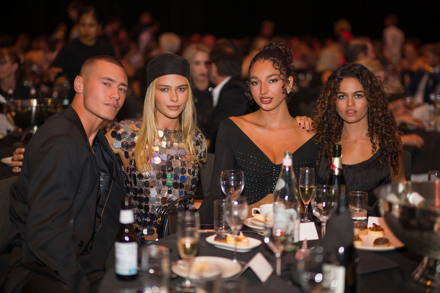 Four guests in evening attire seated at a formal dinner table with wine glasses.