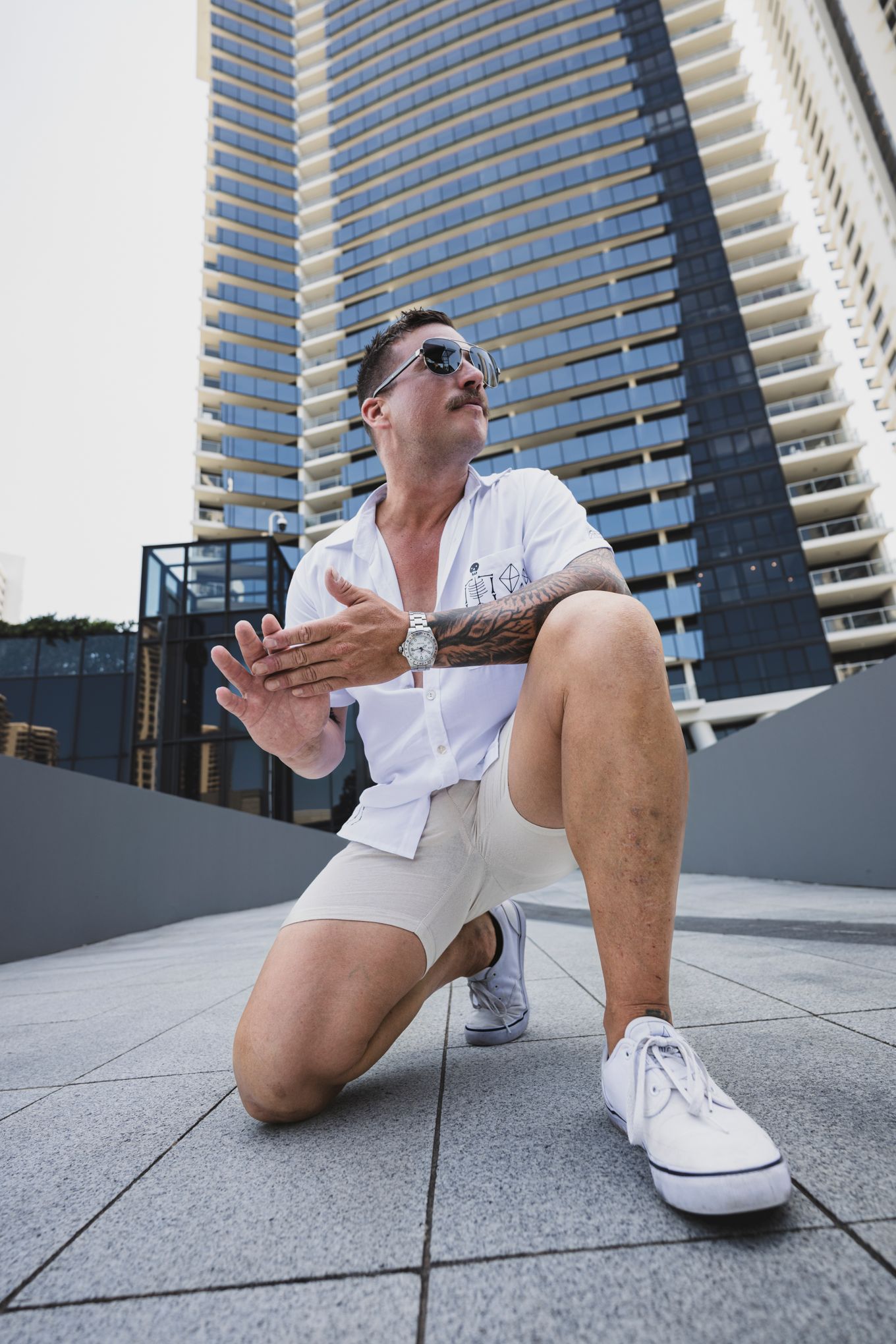 Person in white sneakers and shorts sitting on concrete surface against modern high-rise building.