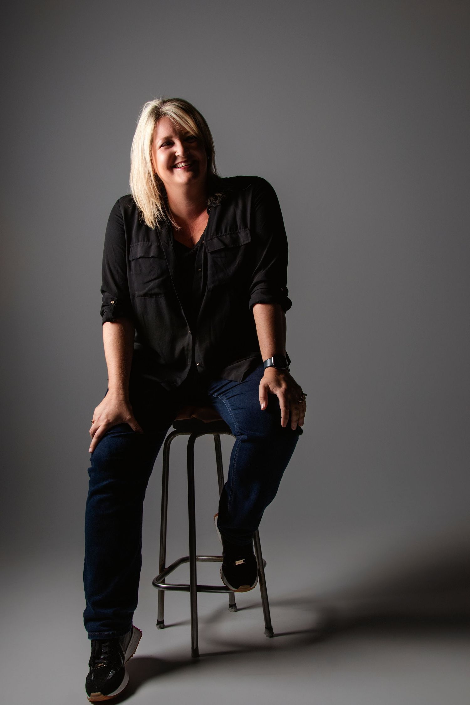 Dramatic studio portraits on stool against dark grey background.