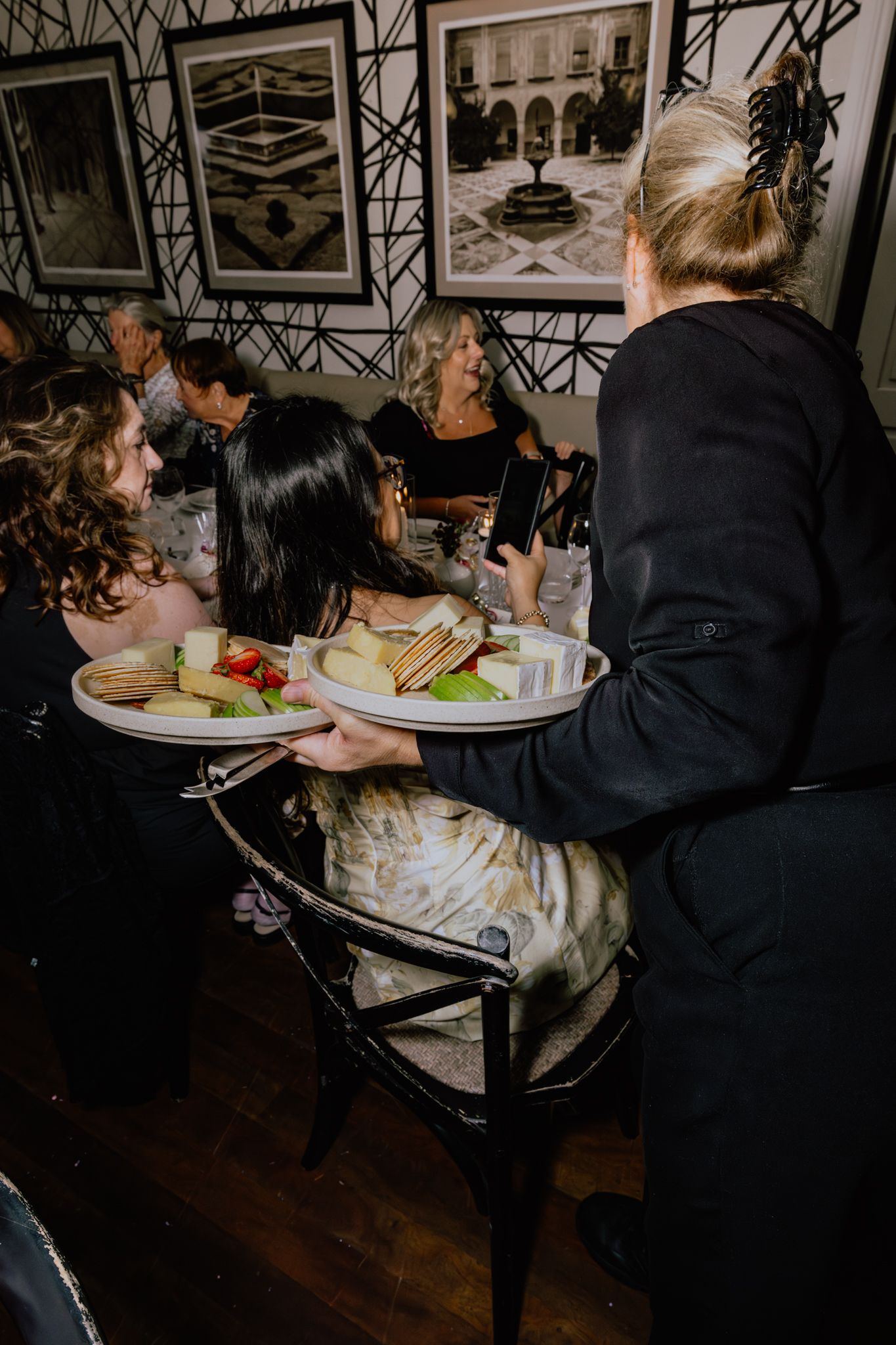 Server carrying plates of food to diners seated at a restaurant with framed artwork on geometric patterned walls.
