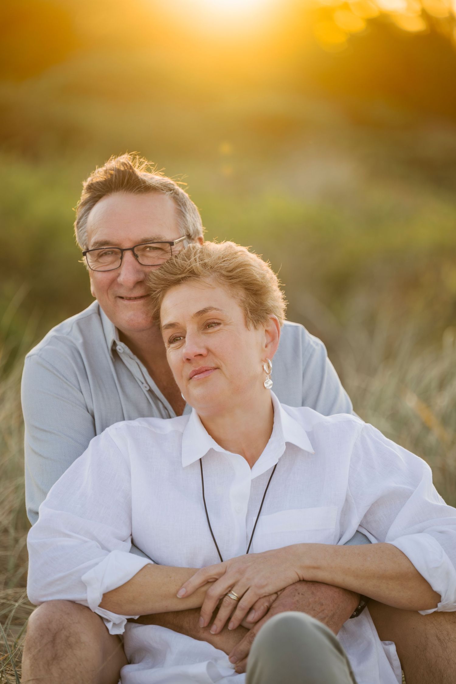 Intimate couple embracing in golden sunset light while sitting in grassy field.