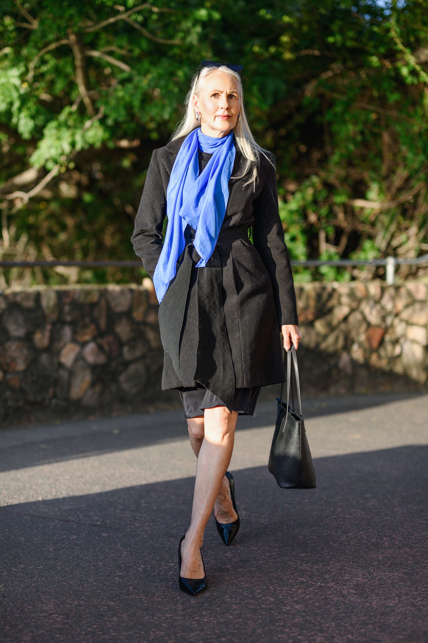Professional look with black blazer and blue scarf walking against stone wall backdrop.