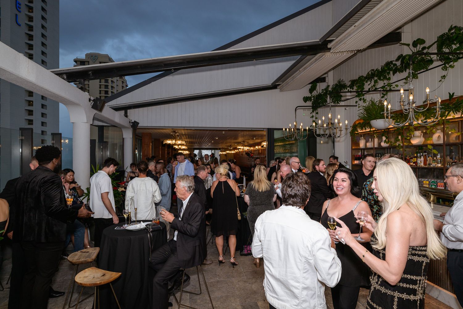 Crowded evening social event on rooftop venue with guests mingling at bar.