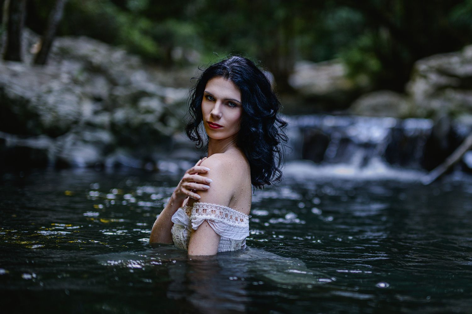 A dark-haired woman in a white off-shoulder dress stands waist-deep in a serene forest river near a waterfall. Cougal cascades, Currumbin