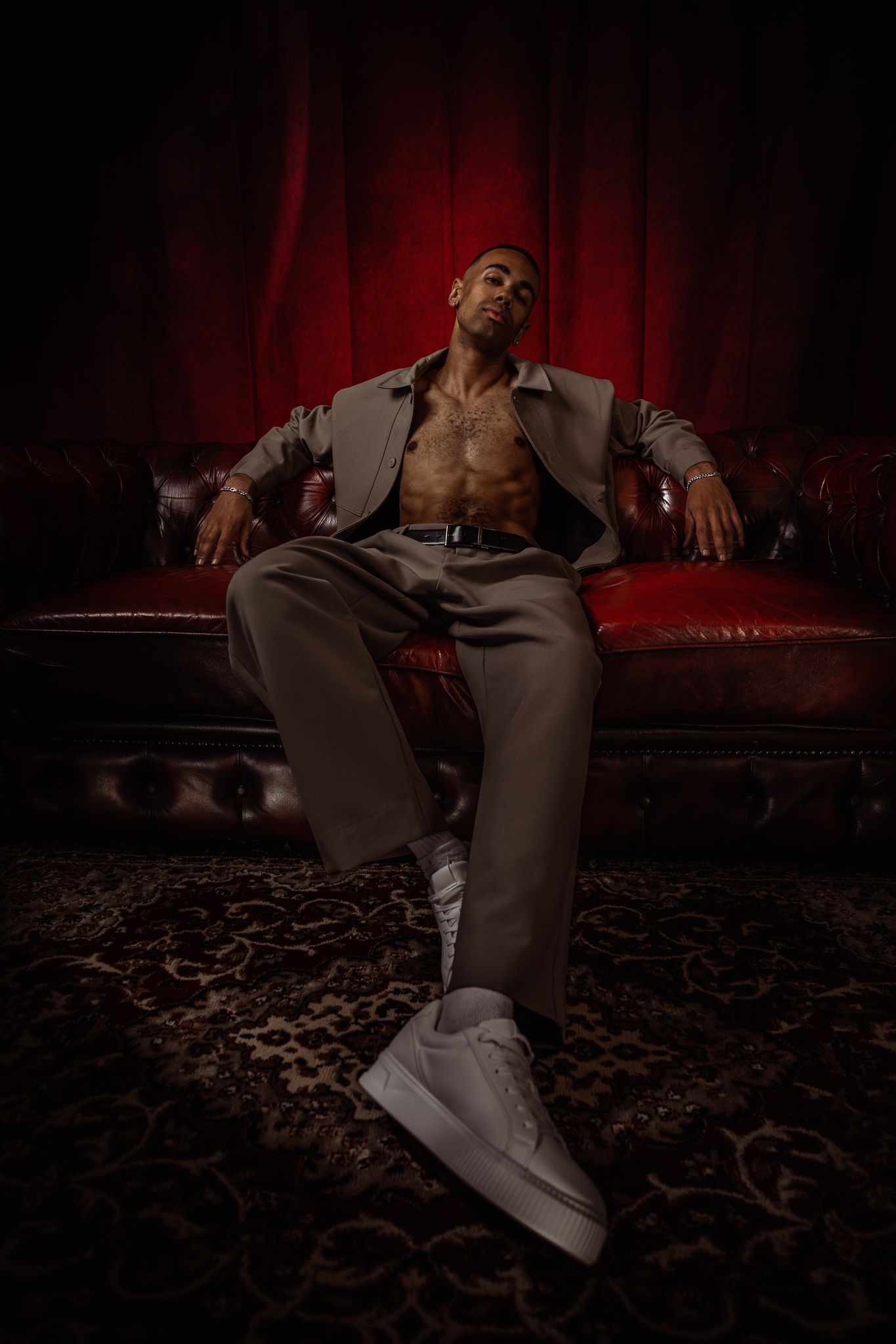 A confident man in an open gray suit sits on a leather sofa against a dramatic red backdrop.