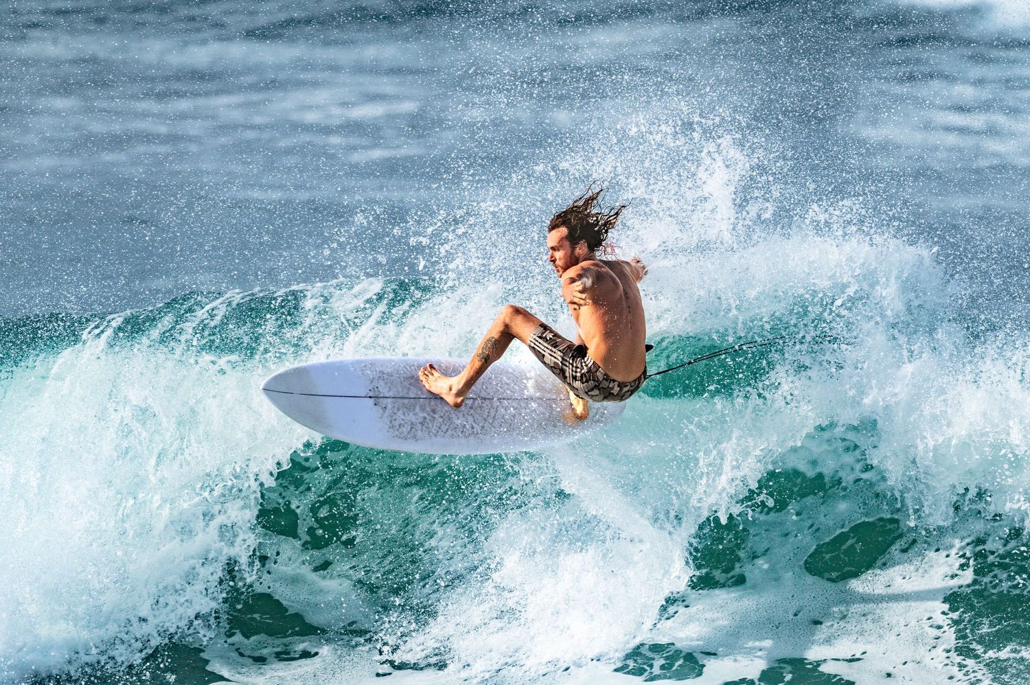 A skilled surfer rides a crashing ocean wave on a white surfboard, with water spraying dramatically around him.