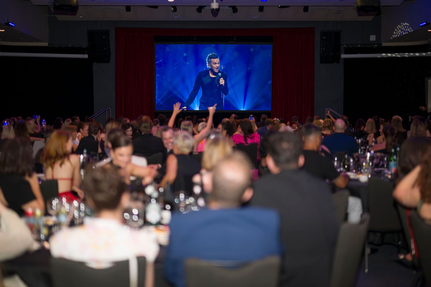 Audience members at round tables watch a performer on a large screen during an indoor event.