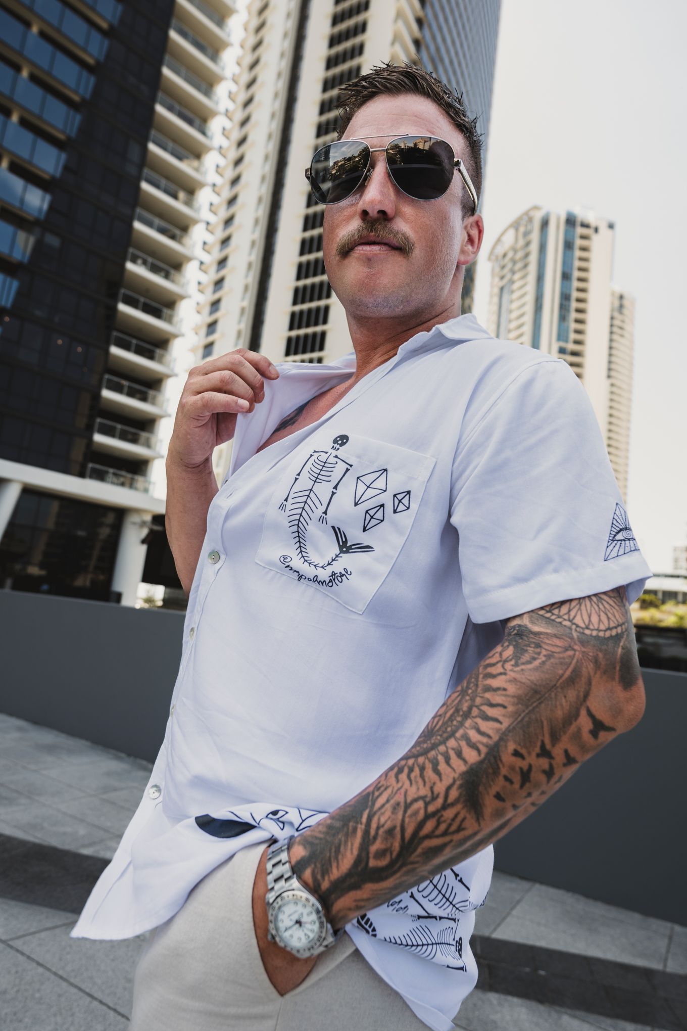 Person wearing white shirt with tattoo sleeve poses against urban backdrop.